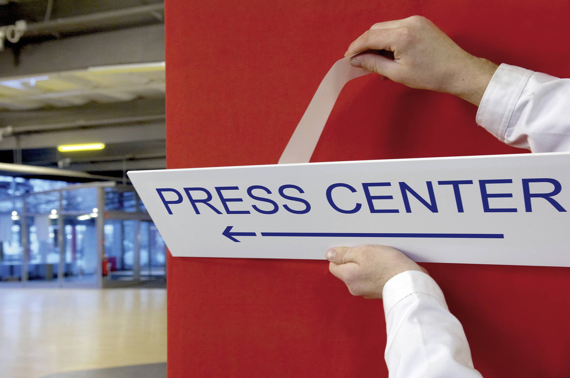 Ein Schild mit der Aufschrift 'PRESS CENTER' wird an einer roten Wand befestigt. Umgebung ist ein geräumiger, modern aussehender Bereich.