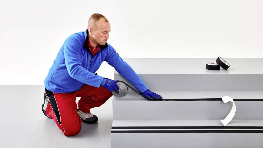Ein Mann kniet und bringt rutschfeste Streifen auf eine Treppenstufe auf. Er trägt Arbeitskleidung und Schutzhandschuhe.