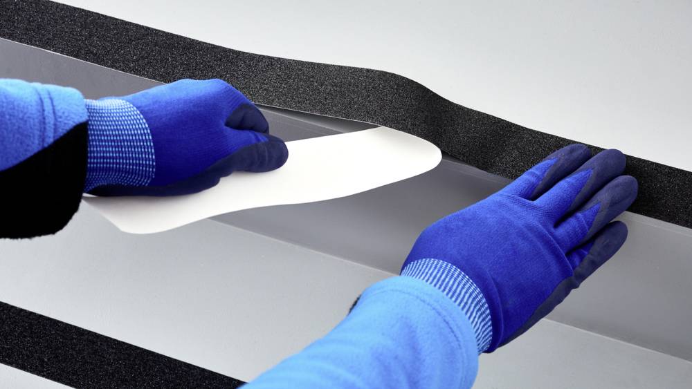 Zwei behandschuhte Hände kleben einen schwarzen Streifen auf eine glatte, graue Oberfläche.