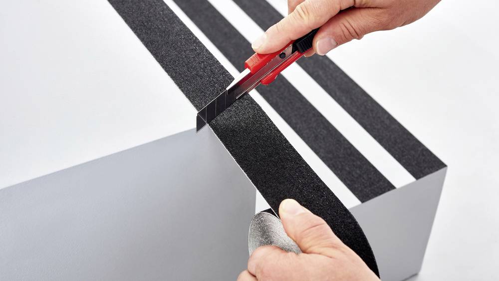 Eine Person schneidet mit einem Cutter-Messer schwarze Anti-Rutsch-Streifen auf eine weiße Oberfläche.