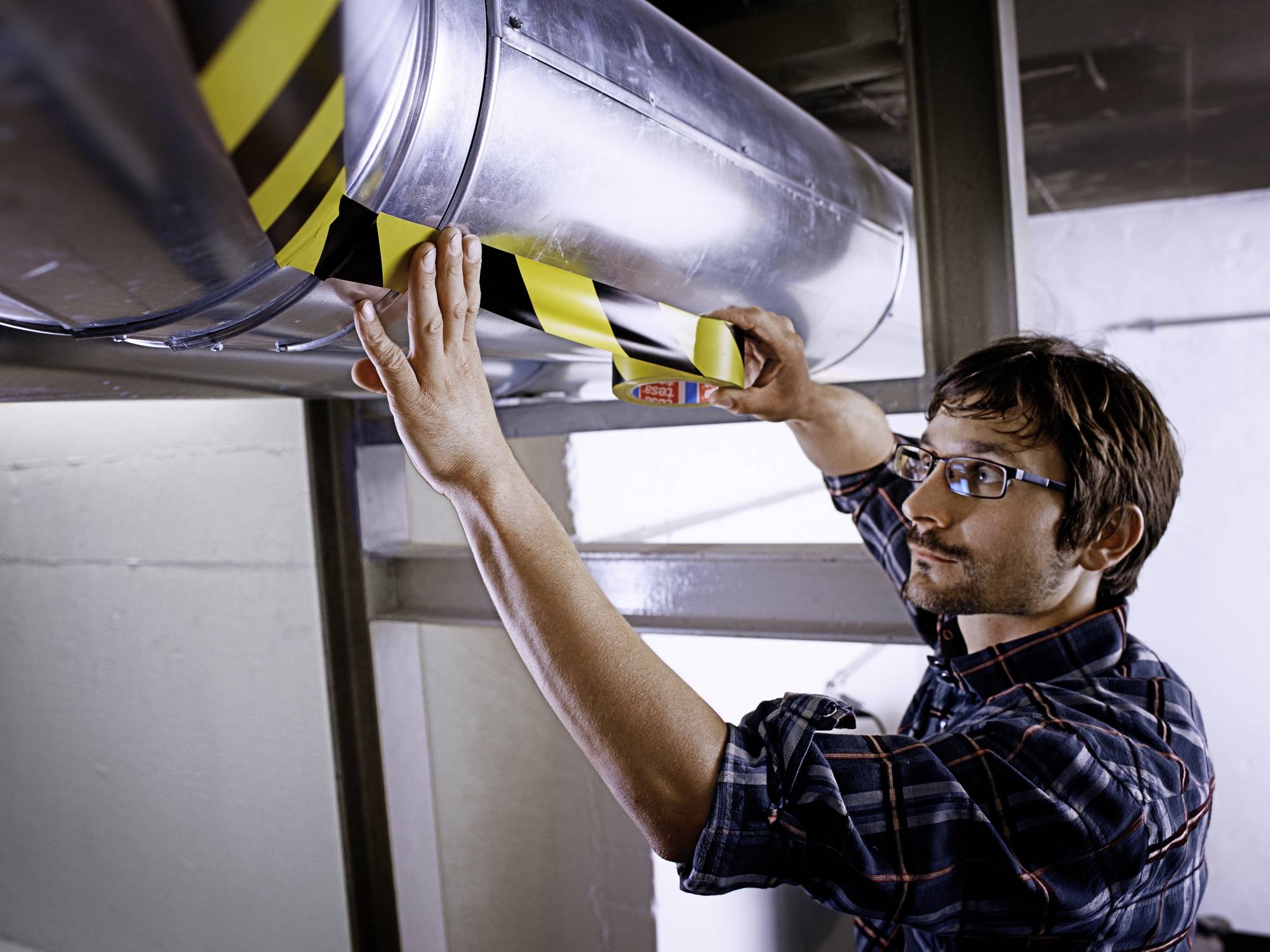 Ein Mann überprüft ein Lüftungsrohr und klebt gelbes Warnband daran. Er trägt eine Brille und ein kariertes Hemd.