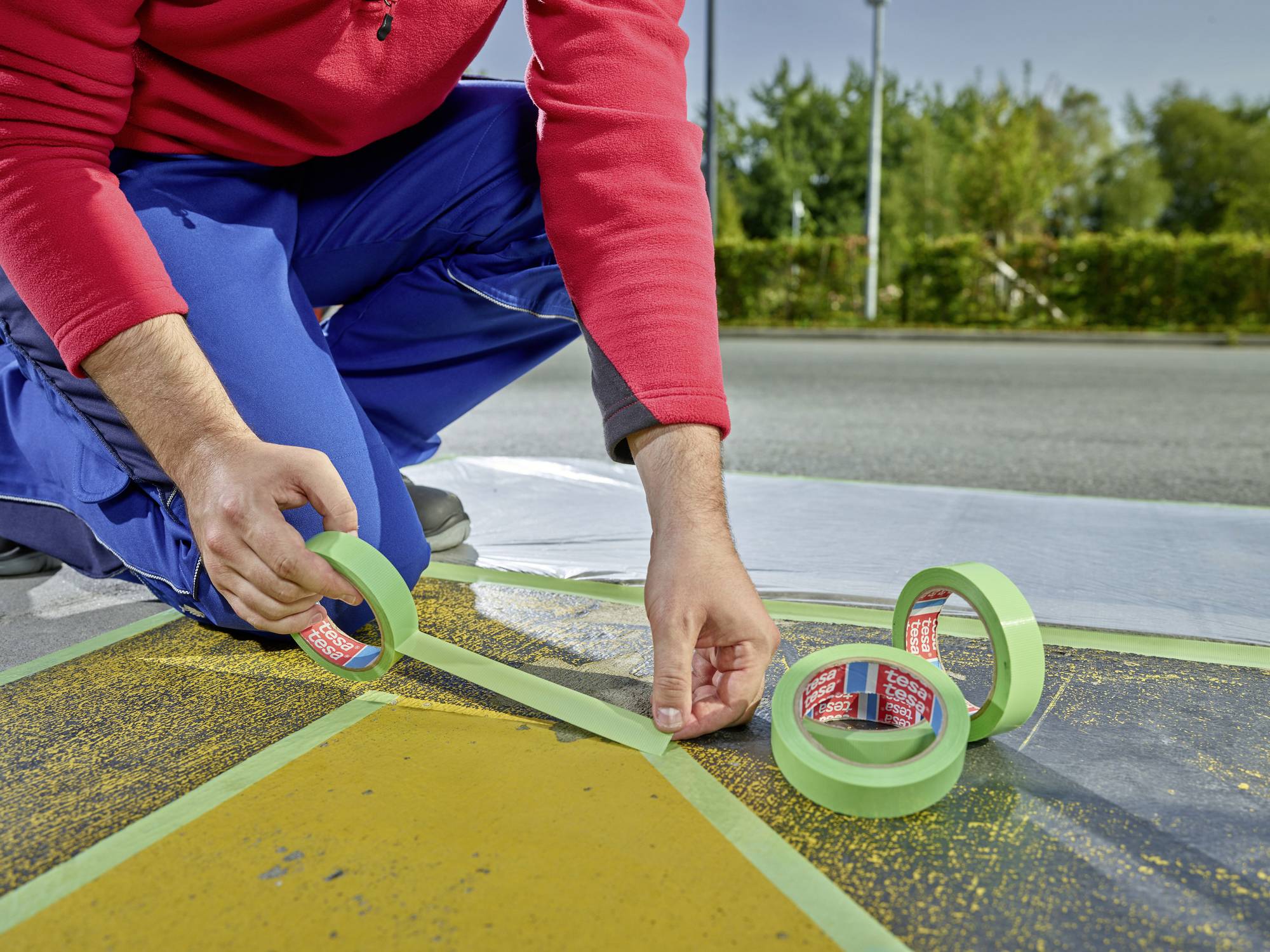 Eine Person kniet auf einer Straße und klebt grünes Klebeband entlang gelber Linien, wahrscheinlich um Markierungen zu erstellen.