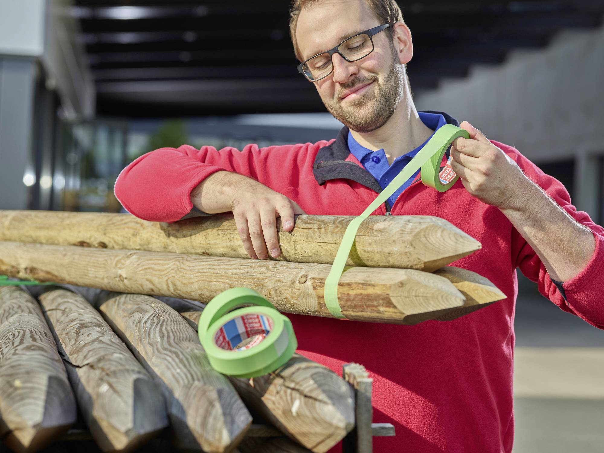 Ein Mann mit Brille und rotem Pullover befestigt Holzpfähle mit grünem Klebeband. Im Hintergrund sind Gebäude erkennbar.