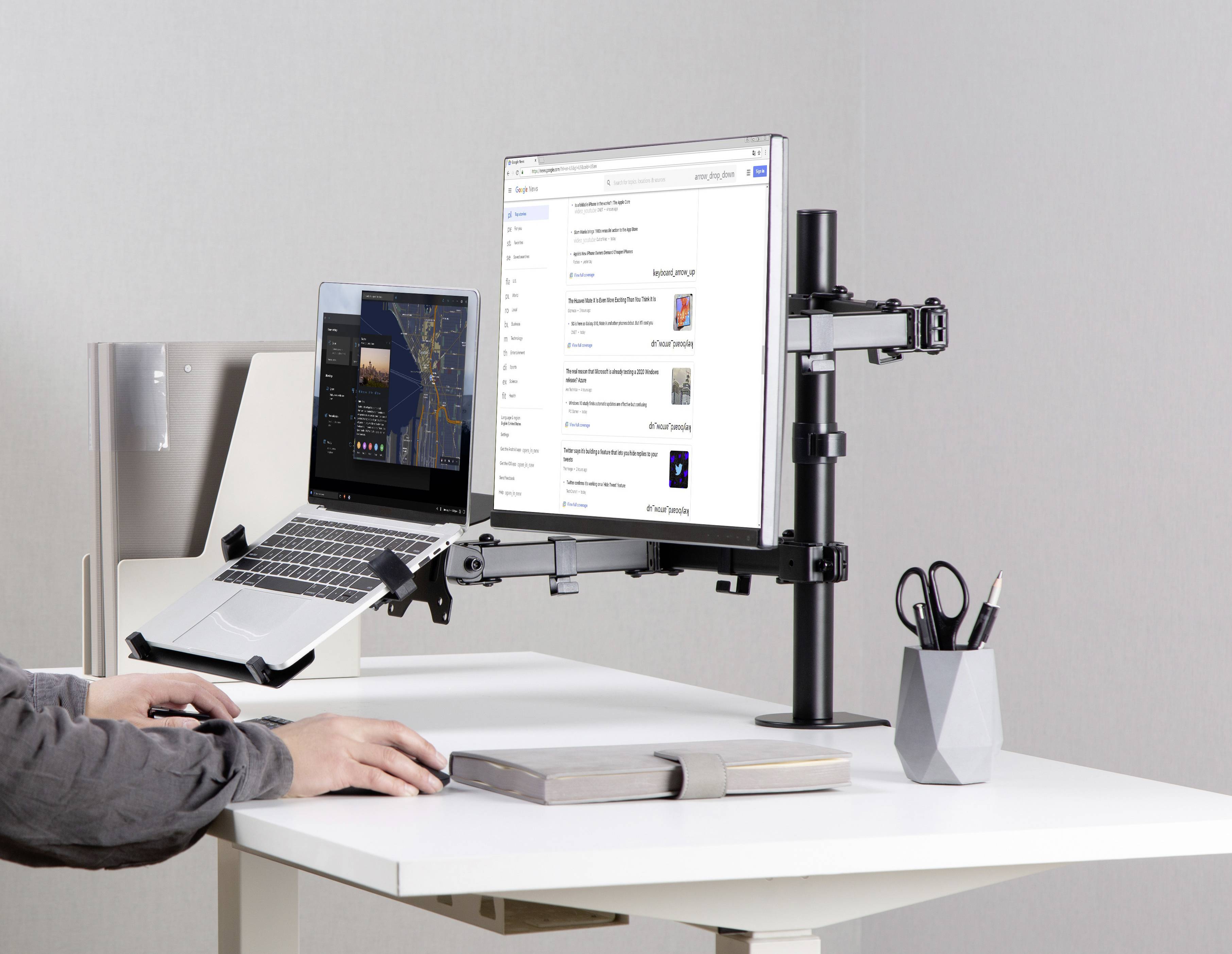 Person arbeitet an einem Schreibtisch mit einem Laptop und einem Monitor, die auf verstellbaren Halterungen stehen. Daneben Stiftehalter und Bücher.