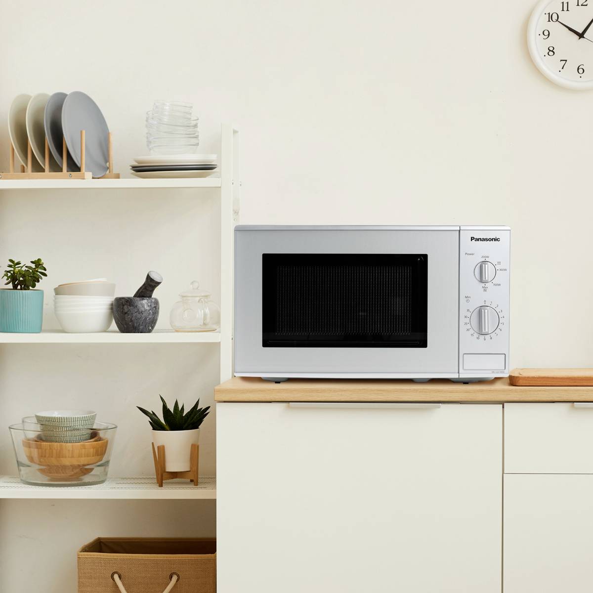 Eine Mikrowelle steht auf einer Küchenzeile. Links befinden sich Regale mit Geschirr und Pflanzen. Rechts ist eine Uhr zu sehen.