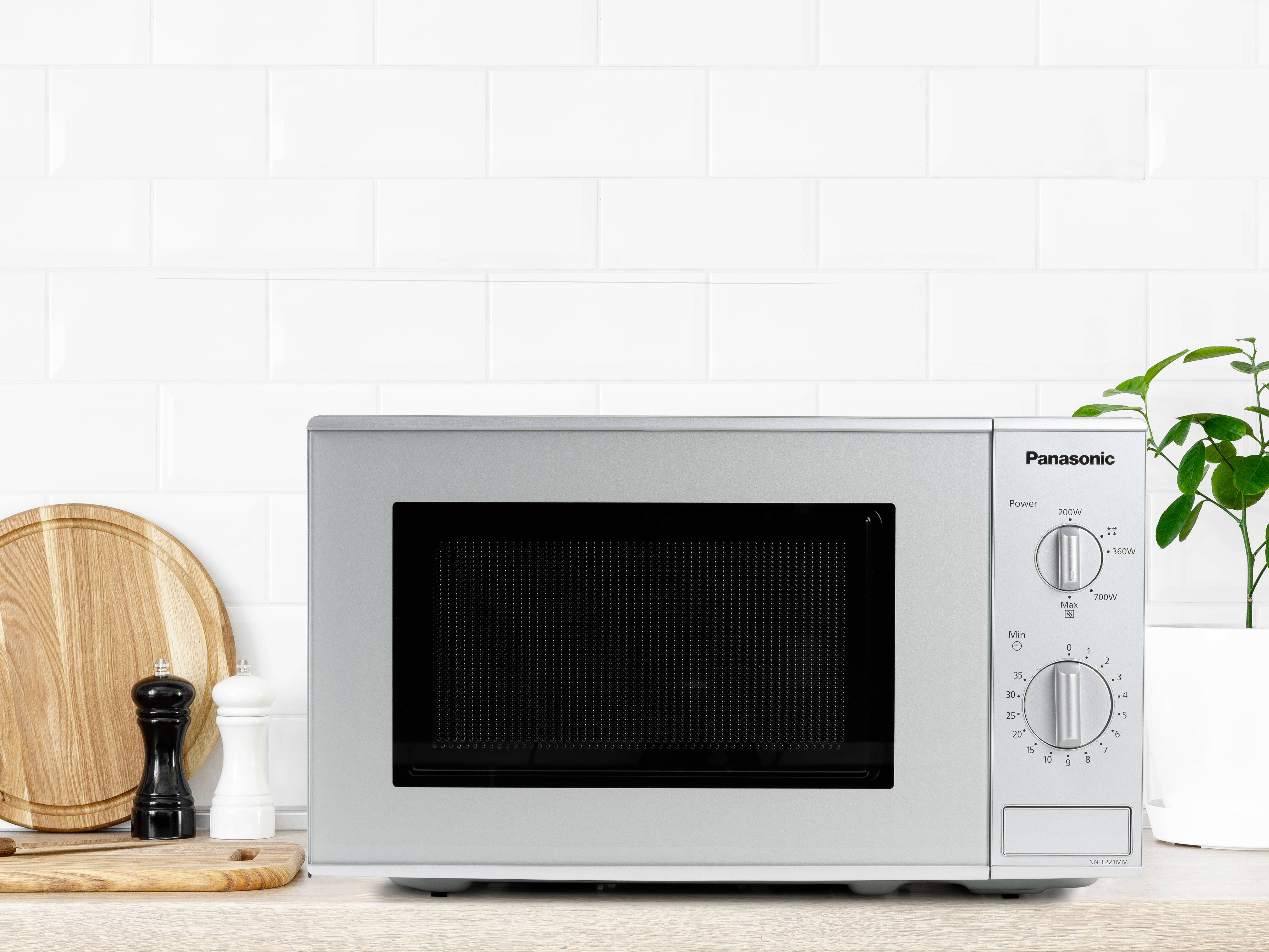 Mikrowelle auf Küchentheke, umgeben von Holzschneidebrett und Gewürzmühlen, vor weißer Fliesenwand.