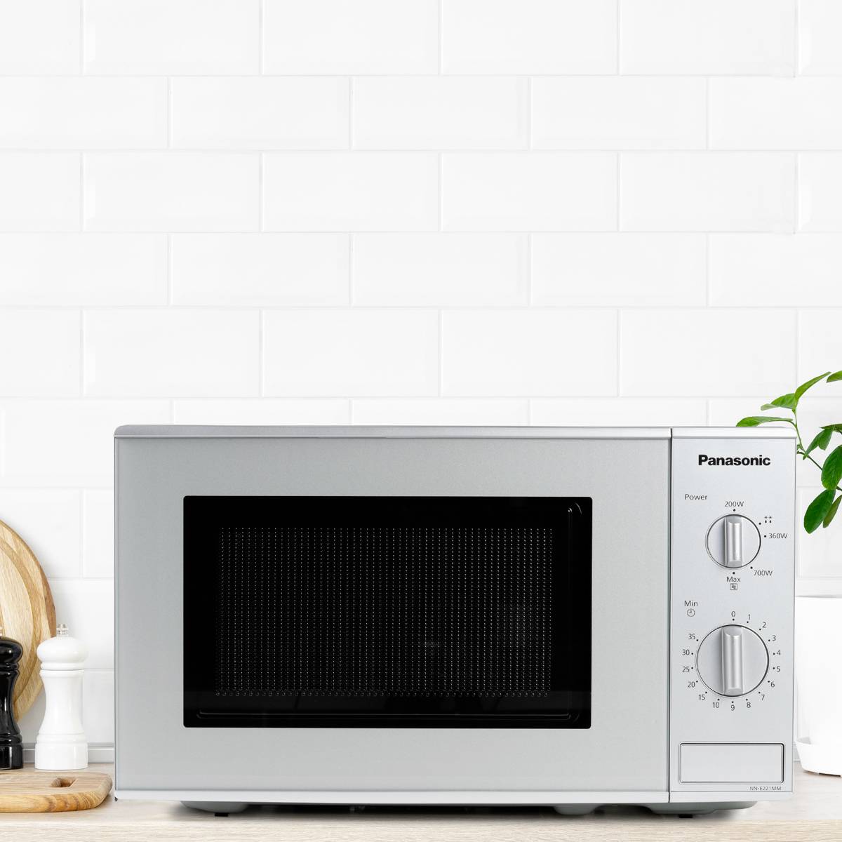 Mikrowelle auf Küchenarbeitsplatte, umgeben von Utensilien. Eierbecher und Schneidebrett links, Pflanze rechts. Schlichtes Küchendesign.