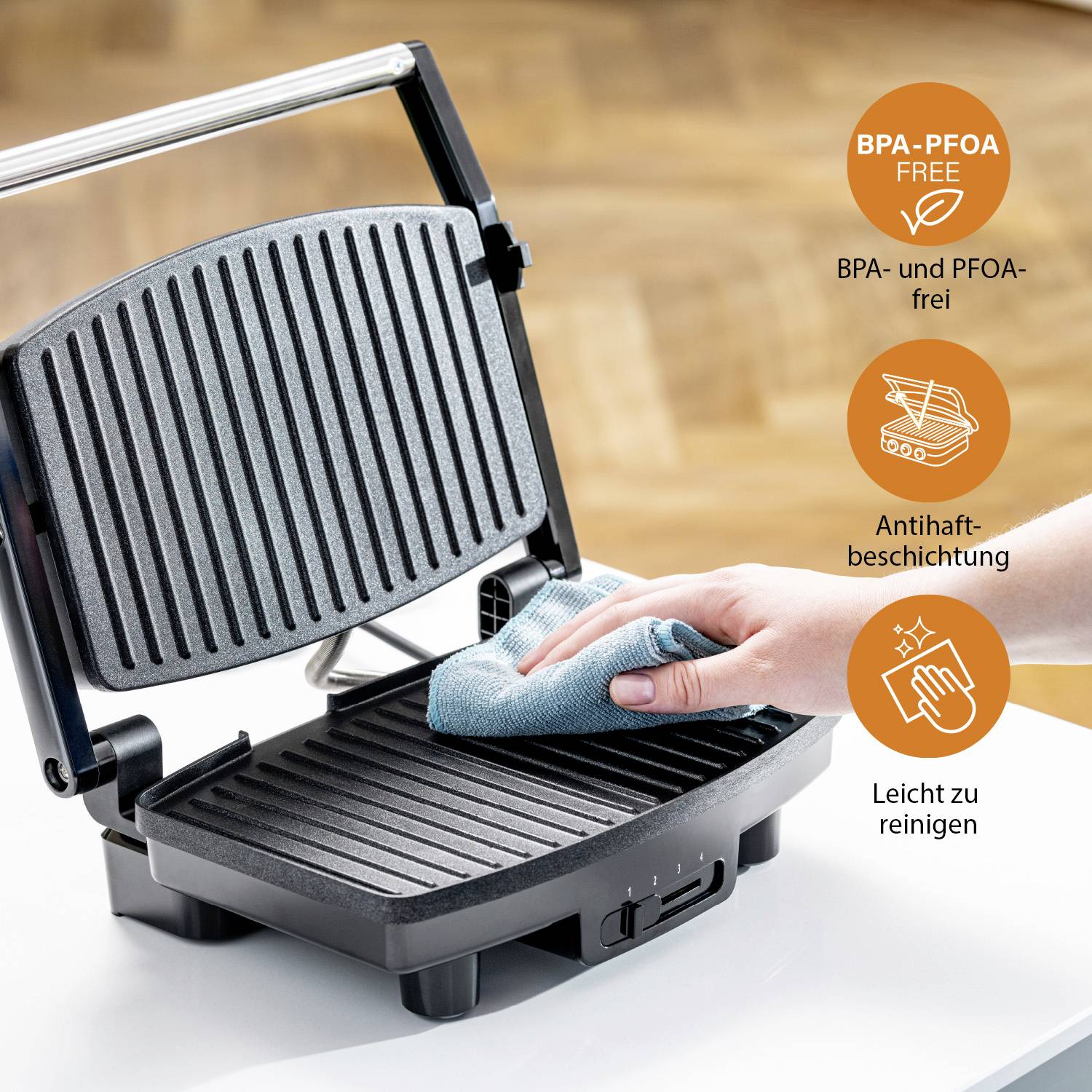 Ein elektrischer Grill wird mit einem Tuch gereinigt. Text: 'BPA- und PFOA-frei, Antihaftbeschichtung, Leicht zu reinigen'.
