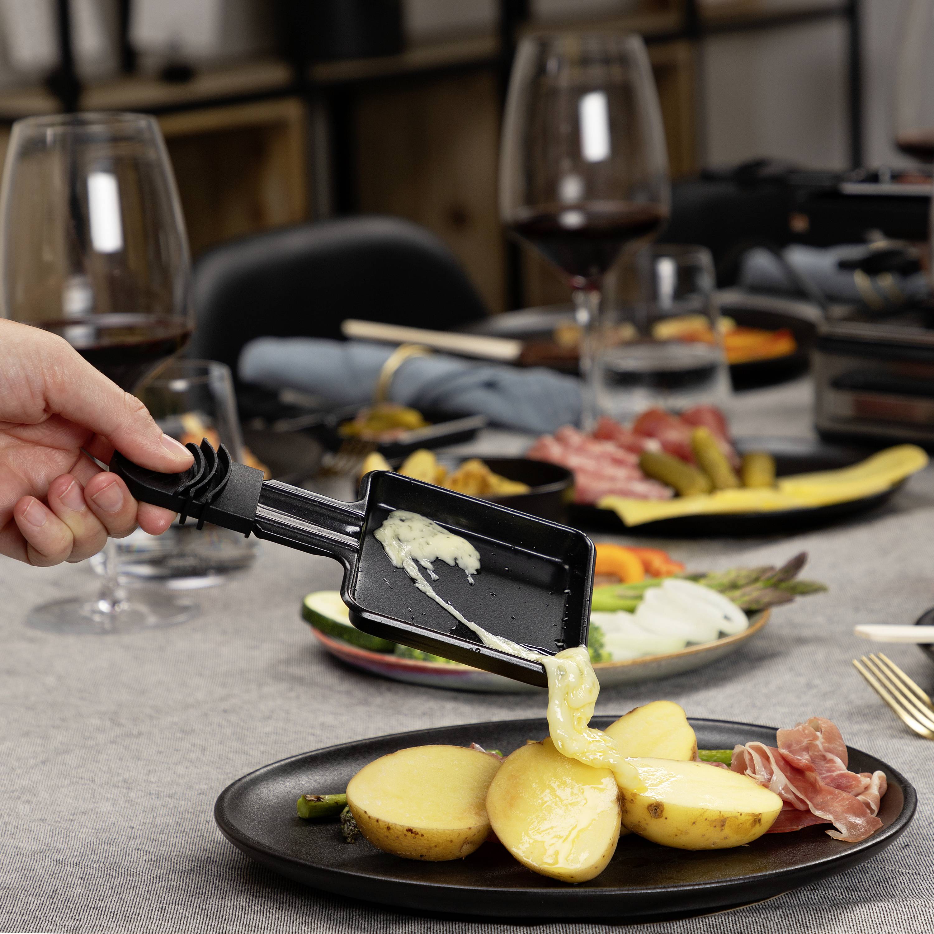 Eine Hand gießt geschmolzenen Käse aus einem Raclette-Pfännchen über Kartoffeln auf einem Teller. Im Hintergrund Wein und Beilagen.