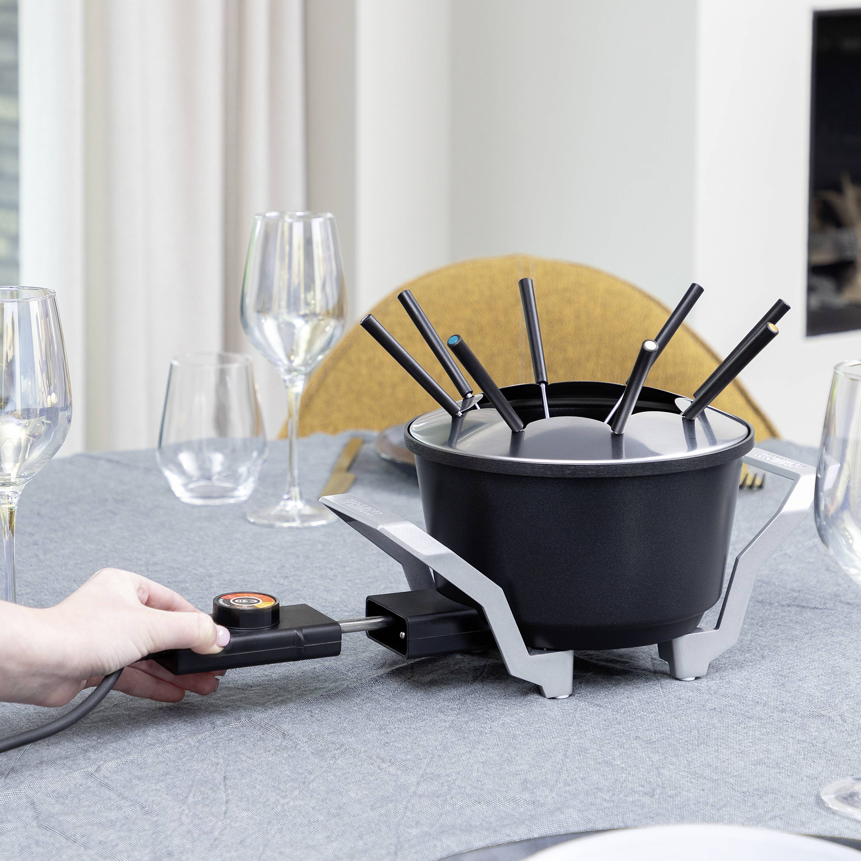 Person richtet einen elektrischen Fonduetopf auf einem gedeckten Tisch her, umgeben von Weingläsern und Tellern.