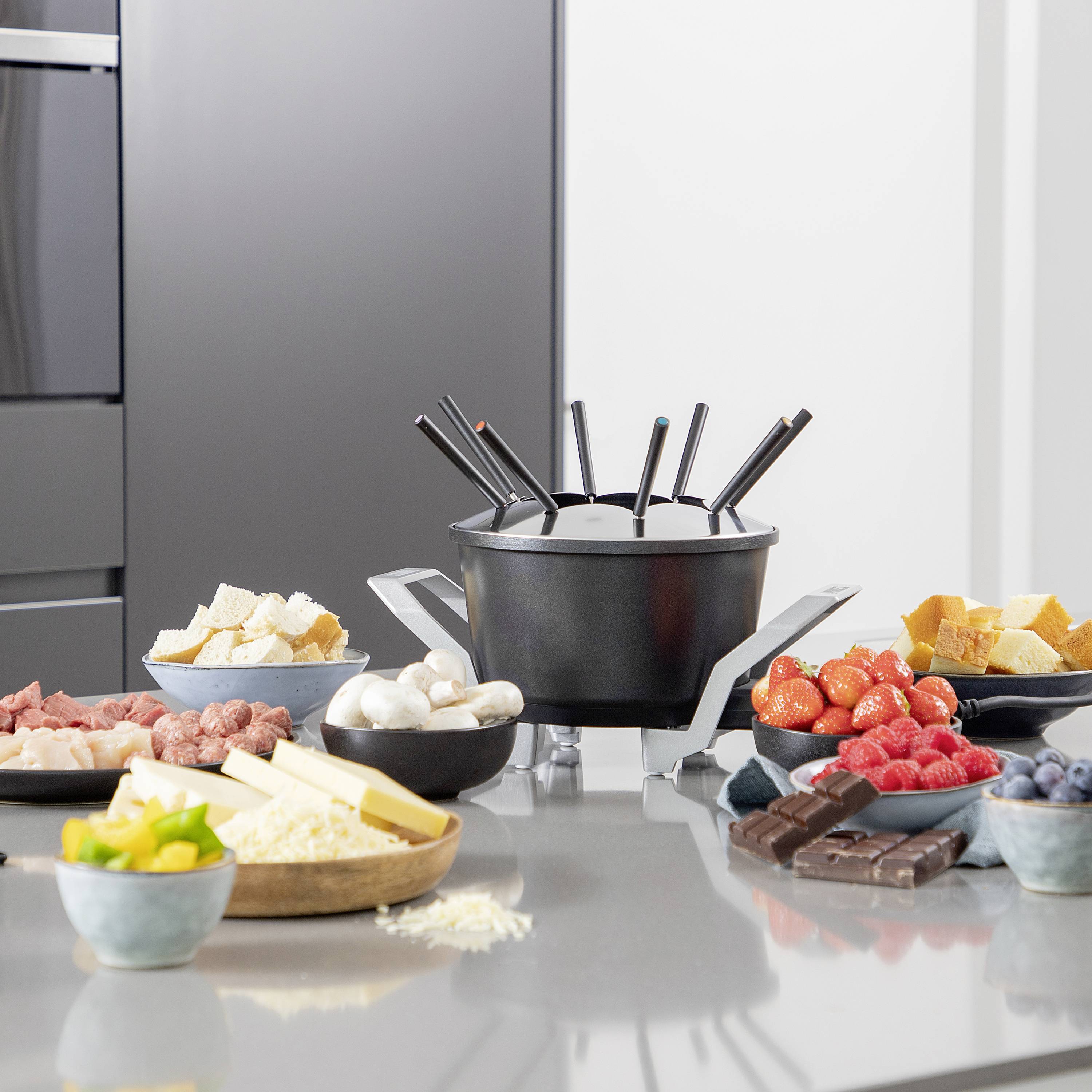 Fondue-Tisch mit erhitztem Topf, umgeben von Fleisch, Gemüse, Brot, Erdbeeren, Heidelbeeren, Schokolade und Käse für eine Mahlzeit.