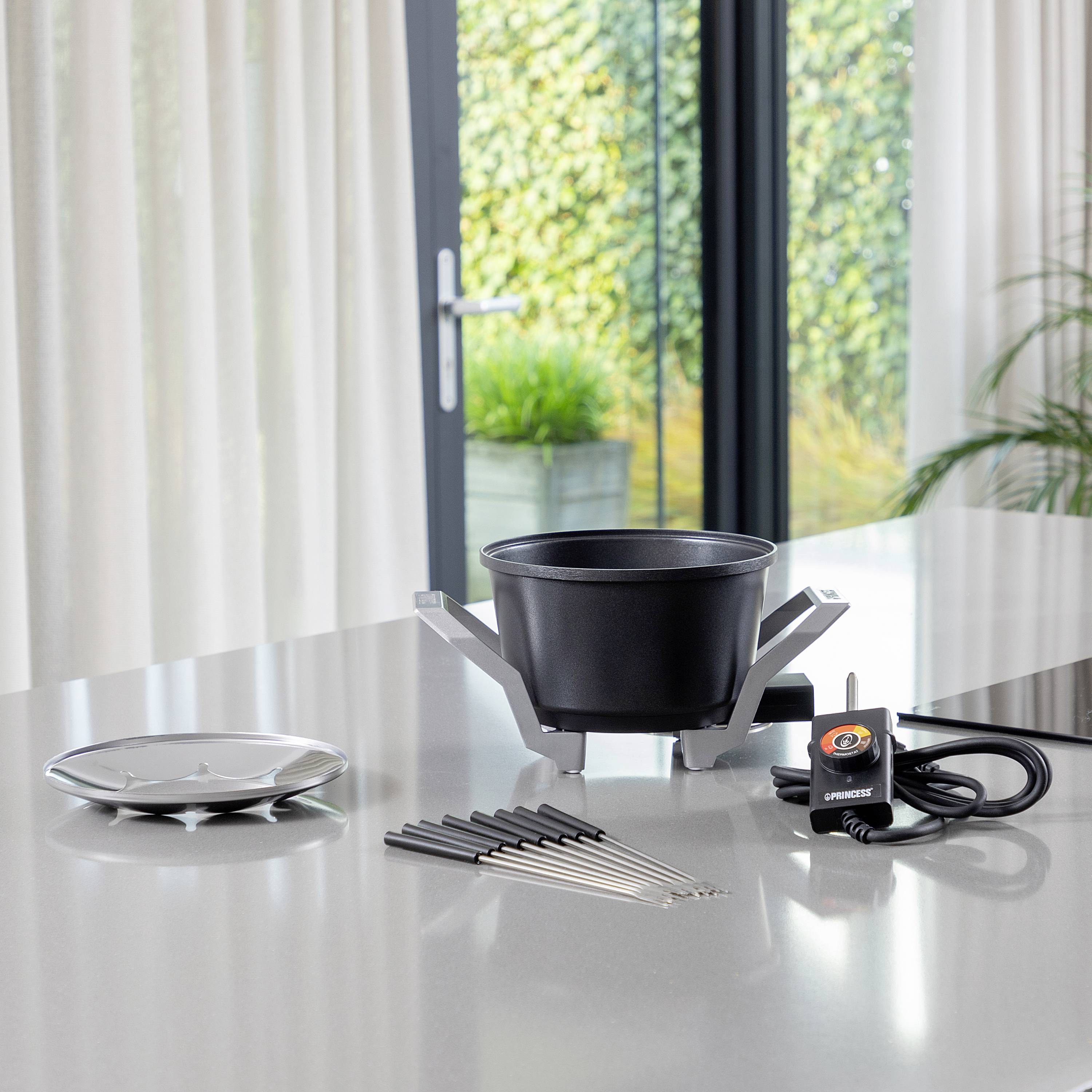 Ein Fonduetopf-Set auf einem Tisch mit Stäbchen, Brenner, und Untersetzer. Im Hintergrund glastür mit Pflanzen außen sichtbar.