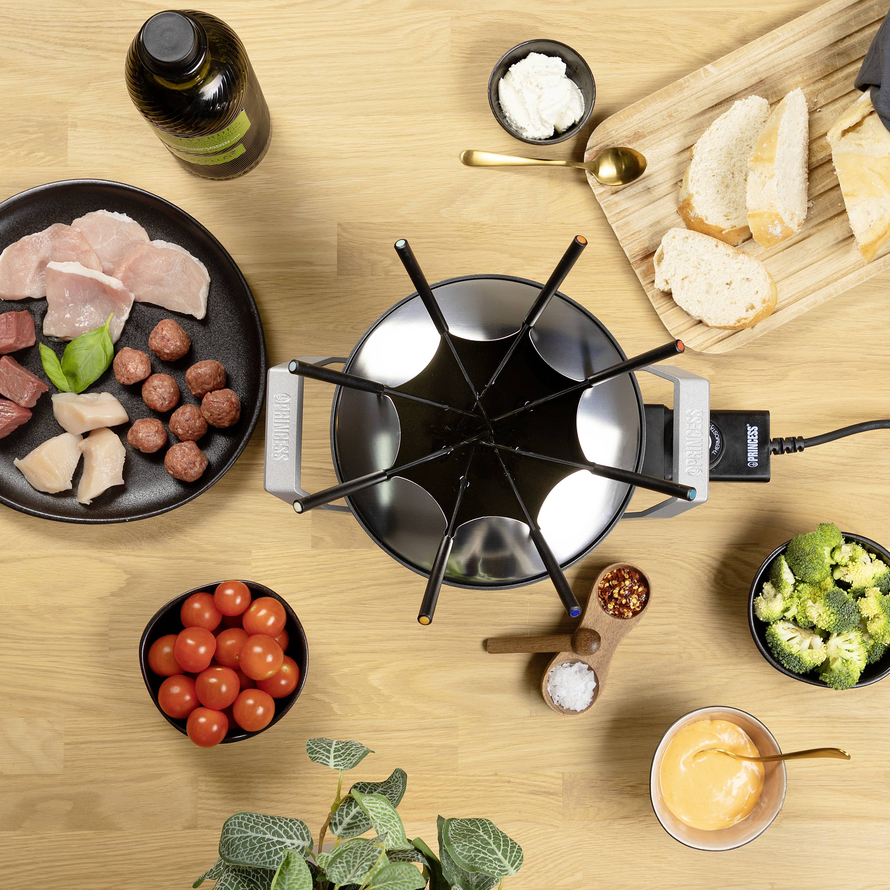 Eine Fondue-Szene mit geschnittenem Fleisch, Brot, Tomaten, Brokkoli und Dips. Ein Fonduetopf steht in der Mitte auf einem Holztisch.