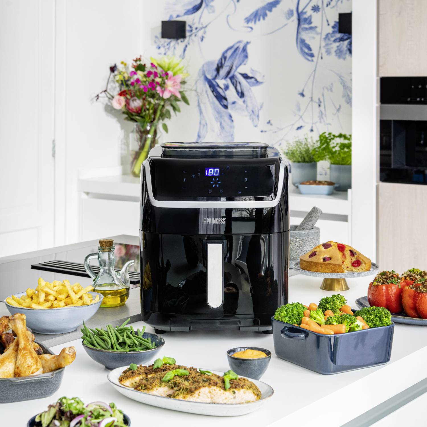 Eine Heißluftfritteuse auf einer Küchenarbeitsplatte, umgeben von verschiedenen Speisen wie Pommes, Hühnchen, gefüllten Paprika und Salat.