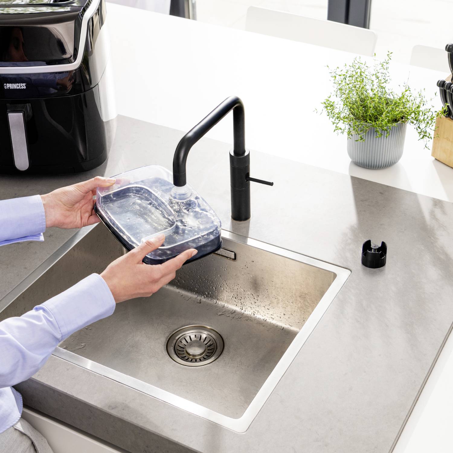 Person füllt Wasserbehälter am Waschbecken auf. Ein Luftreiniger steht auf der Arbeitsplatte. Eine Pflanze steht daneben.