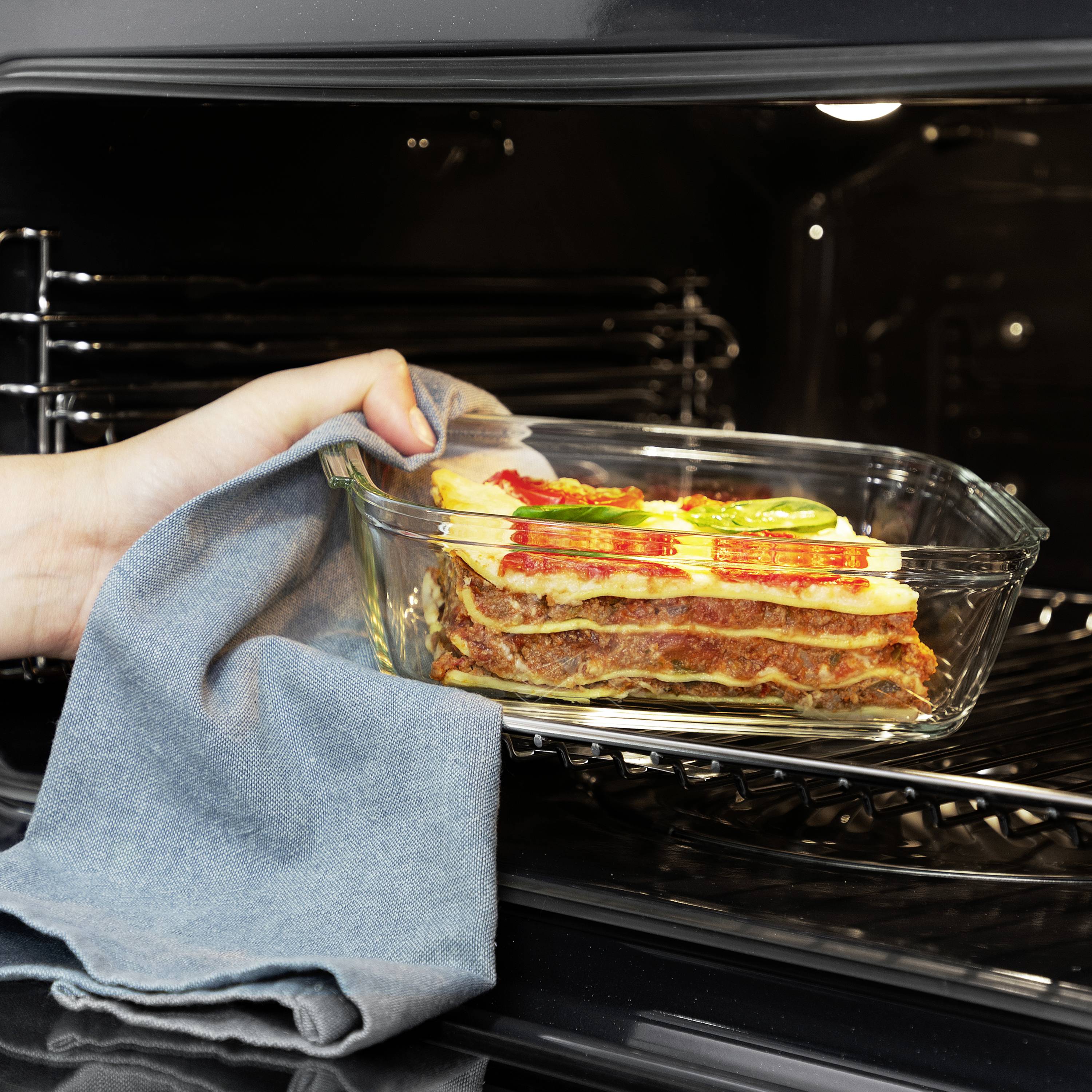 Eine Hand nimmt eine Lasagne in einer durchsichtigen ofenfesten Form aus dem Backofen.
