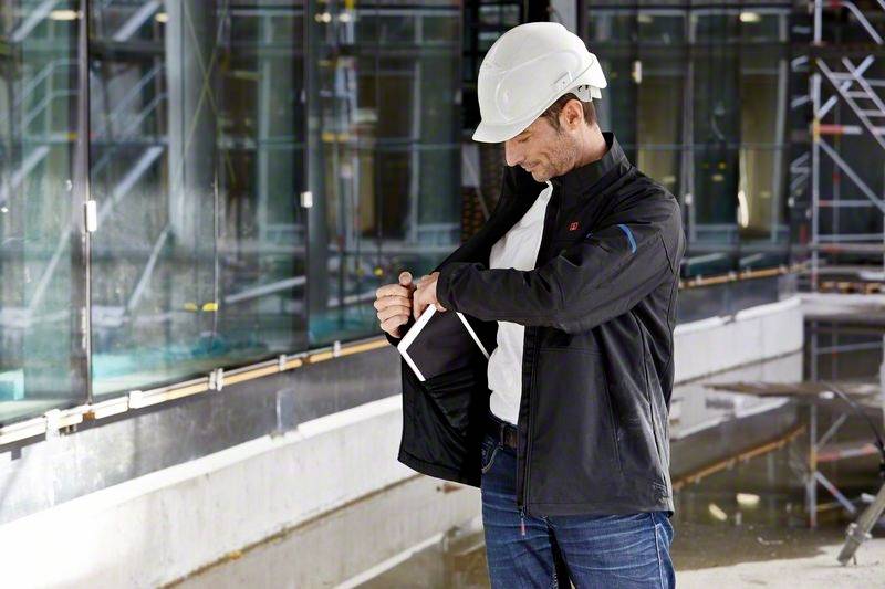 Ein Mann mit Schutzhelm steht auf einer Baustelle und schaut in seine offene Jackentasche, während er ein Tablet hält.