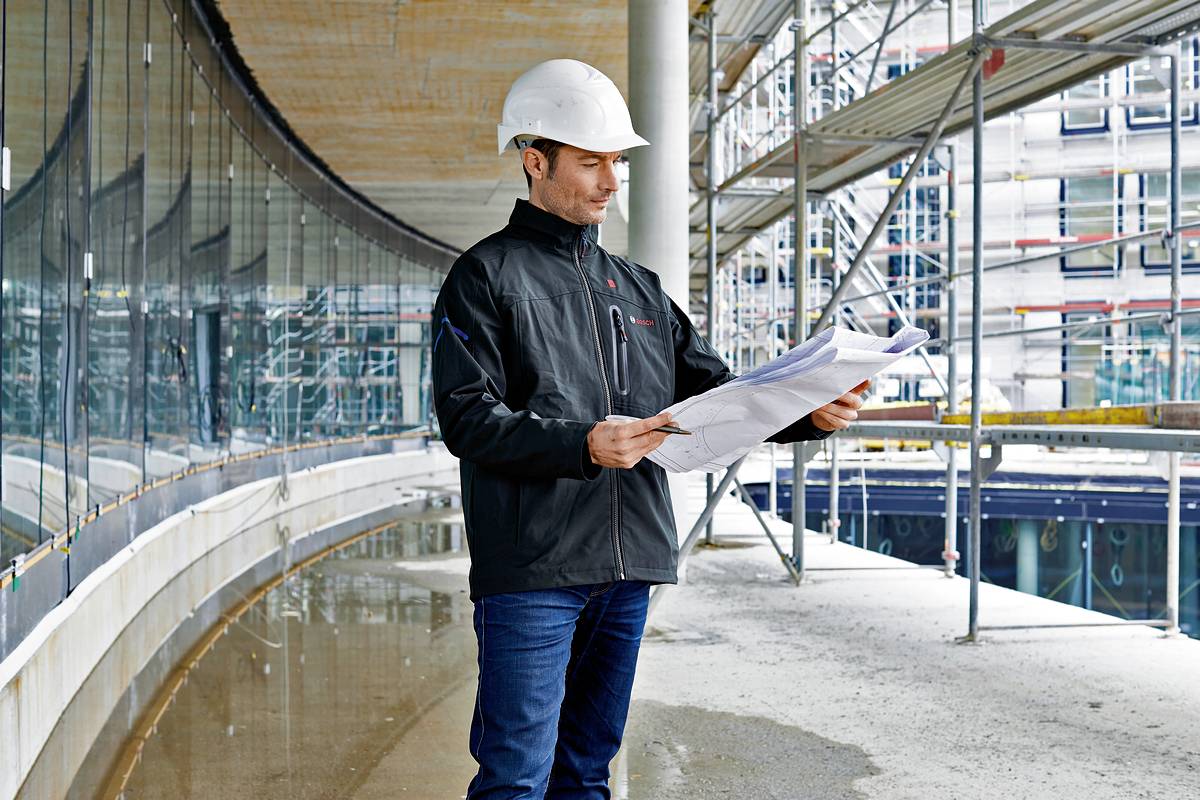 Ein Bauarbeiter mit weißem Schutzhelm betrachtet Baupläne auf einer Baustelle mit Gerüsten und moderner Glasfassade im Hintergrund.