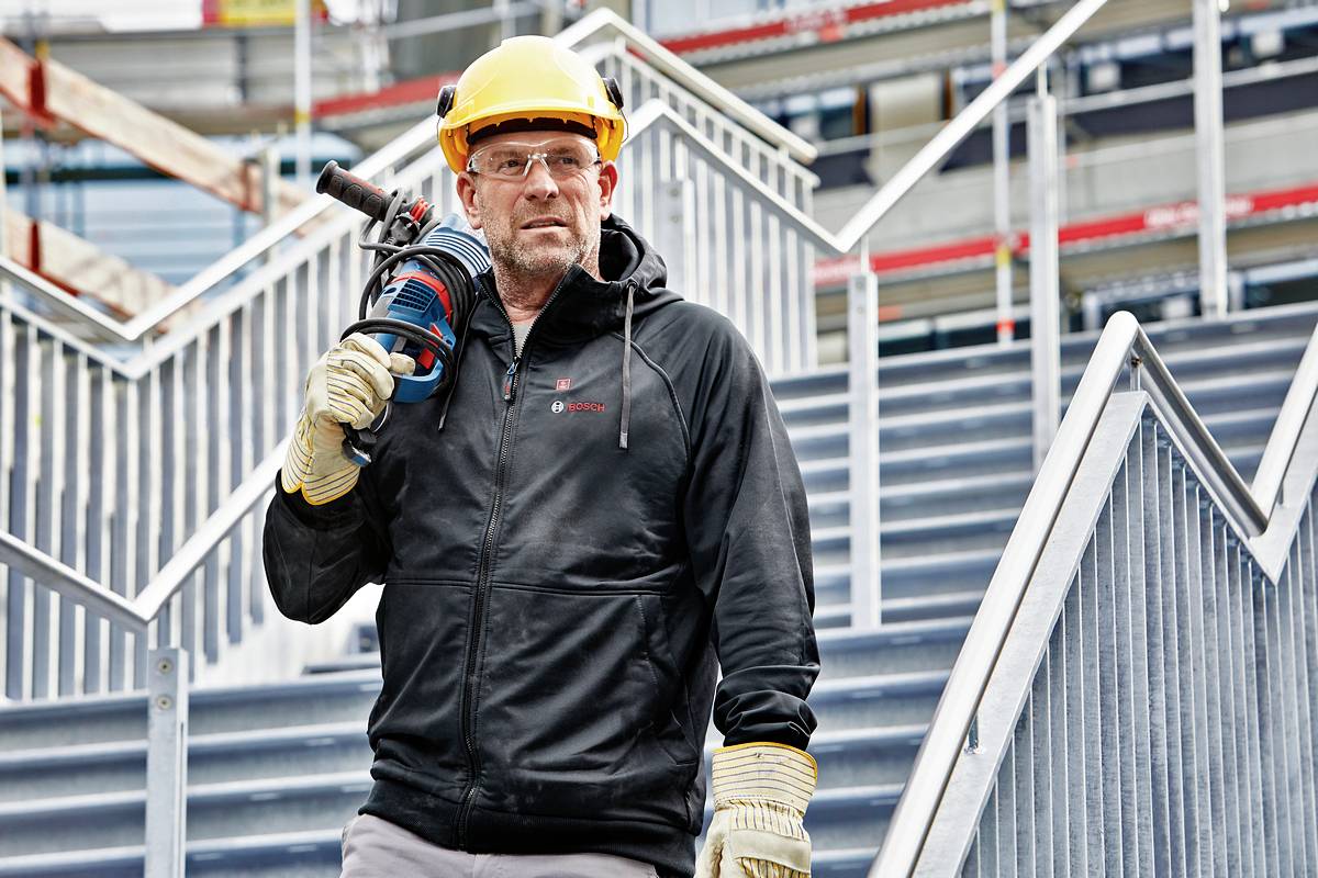 Ein Bauarbeiter mit Helm und Sicherheitskleidung steht auf einer Treppe und hält ein Elektrowerkzeug. Im Hintergrund sind Baugerüste zu sehen.