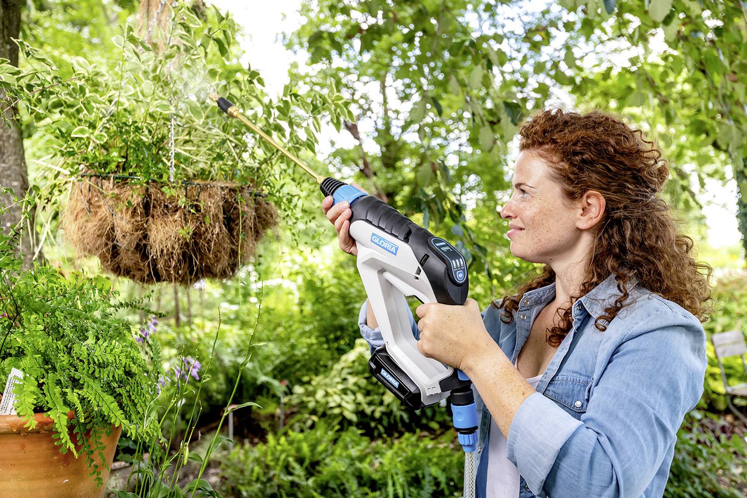 Eine Frau bewässert hängende Pflanzen im Garten mit einem Gartenschlauch. Sie trägt ein Jeanshemd. Umgebung ist grün und blühend.