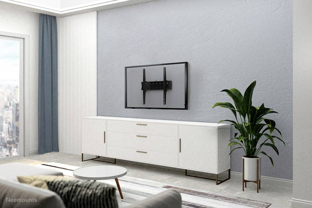 Schlicht eingerichtetes Wohnzimmer mit weißem Sideboard, an der Wand montiertem Fernseher ohne Bildschirm und grüner Pflanze. Im Hintergrund ist eine Stadtansicht aus dem Fenster zu sehen.