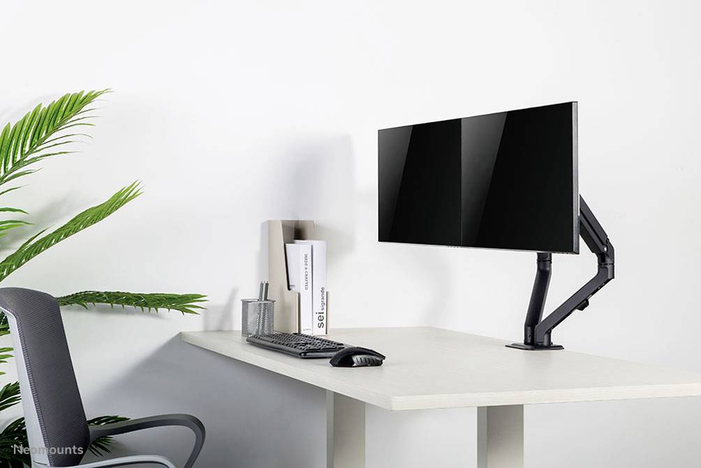 Moderner Schreibtisch mit einem Monitor auf einem beweglichen Arm, Tastatur und Maus. Links eine Topfpflanze und ein Bürostuhl.