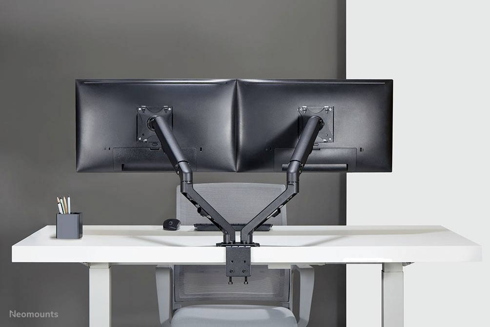 Zwei große Monitore auf einem Schreibtisch an einem doppelten Monitorarm befestigt. Im Hintergrund eine graue Wand.