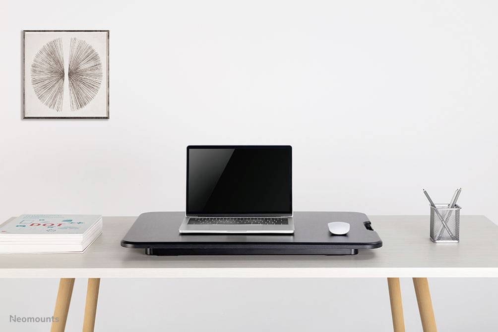 Ein sauberer, moderner Schreibtisch mit einem Laptop auf einem Schwarz-Schreibtischaufsatz, rechts eine Maus, links Bücher. Rechts ein Stiftehalter. Im Hintergrund Kunst an der Wand.