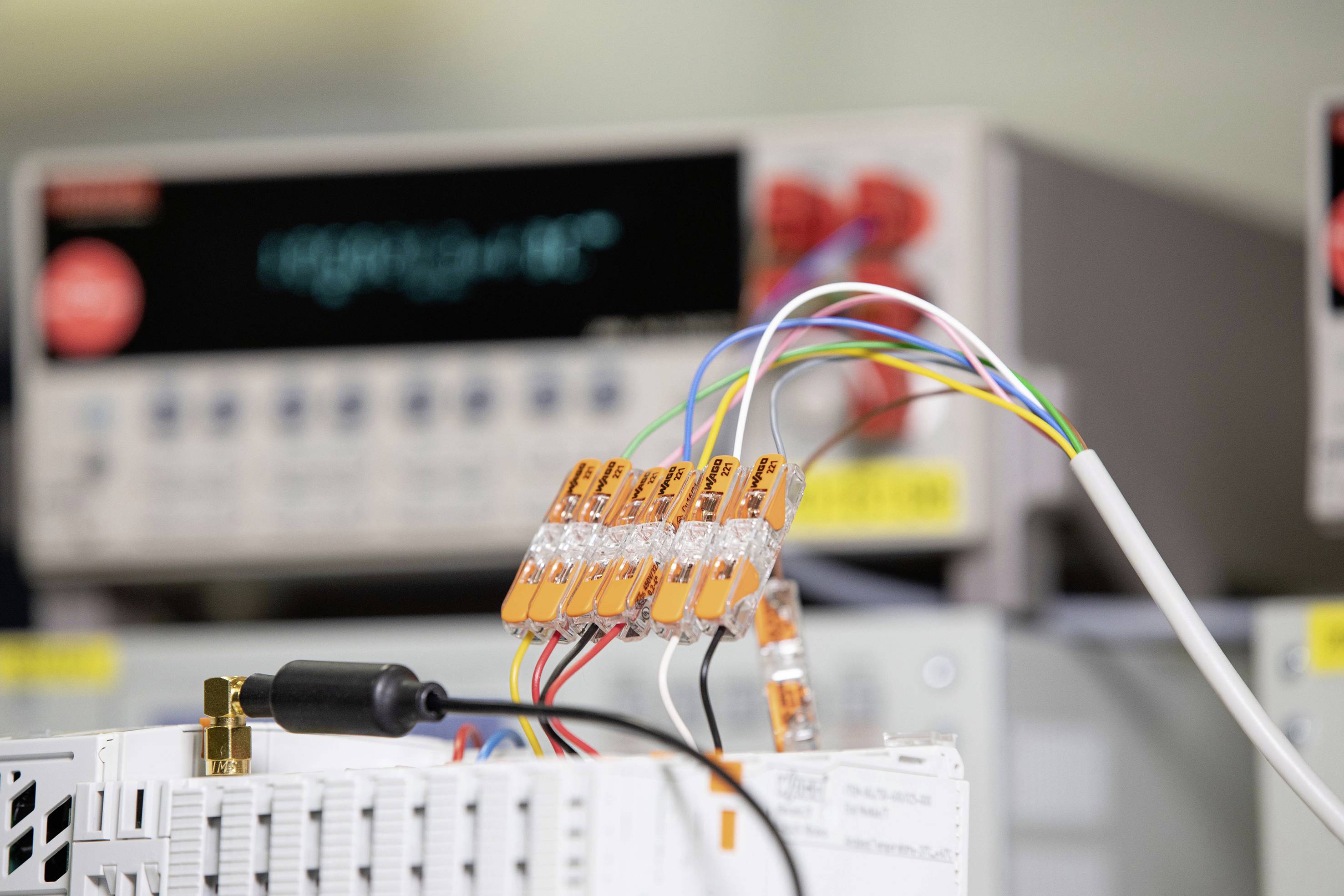 Ein Labor mit elektrischen Messgeräten im Hintergrund. Im Vordergrund sind Kabel mit orangefarbenen Klemmen verbunden.