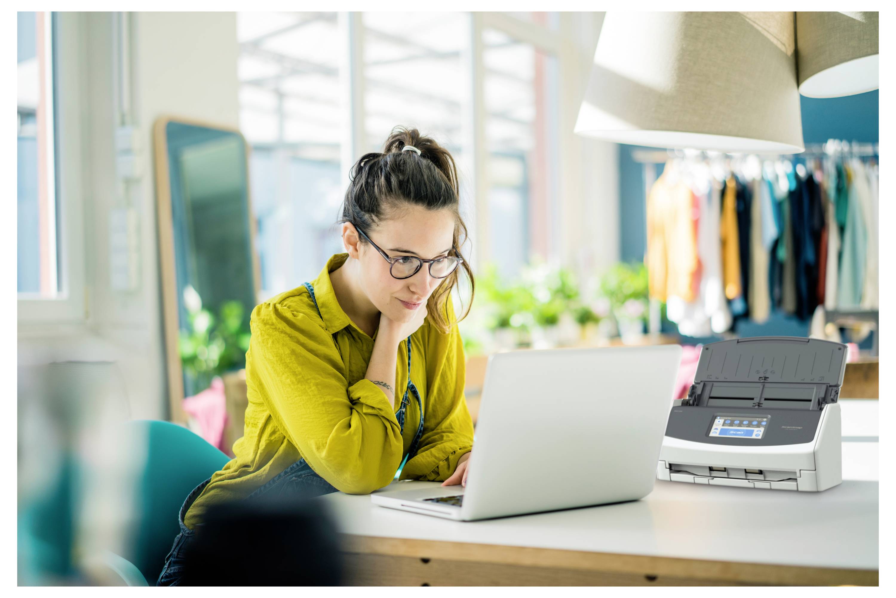 Eine Frau in einem modernen, hell erleuchteten Büro arbeitet an einem Laptop auf einem weißen Schreibtisch. Ein Drucker steht neben ihr. Im Hintergrund sind Kleiderbügel zu sehen.