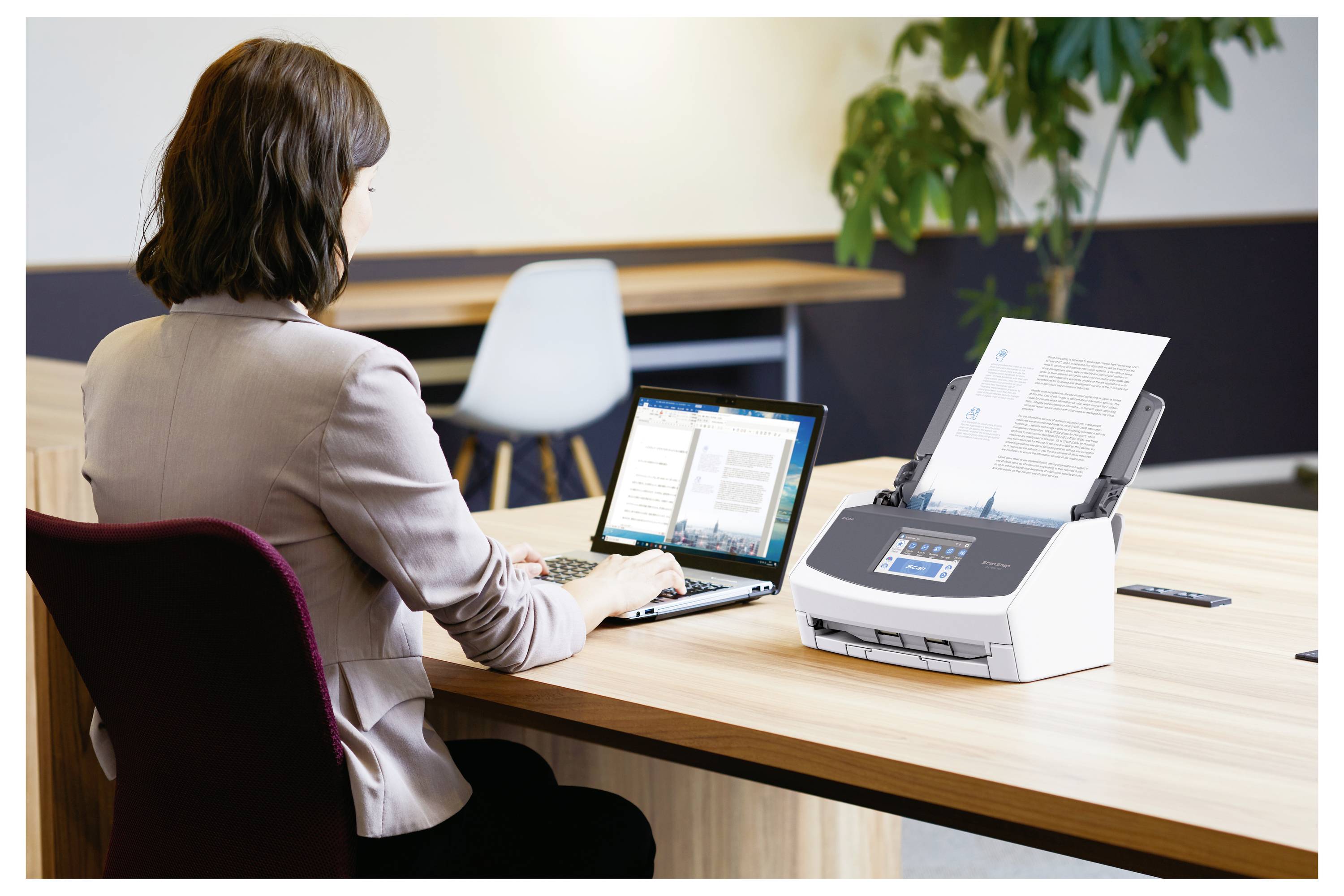 Eine Frau nutzt einen Laptop an einem Schreibtisch, mit einem Einzugscanner rechts von ihr, der gerade Dokumente scannt. Bürosituation mit einem weiteren Schreibtisch.