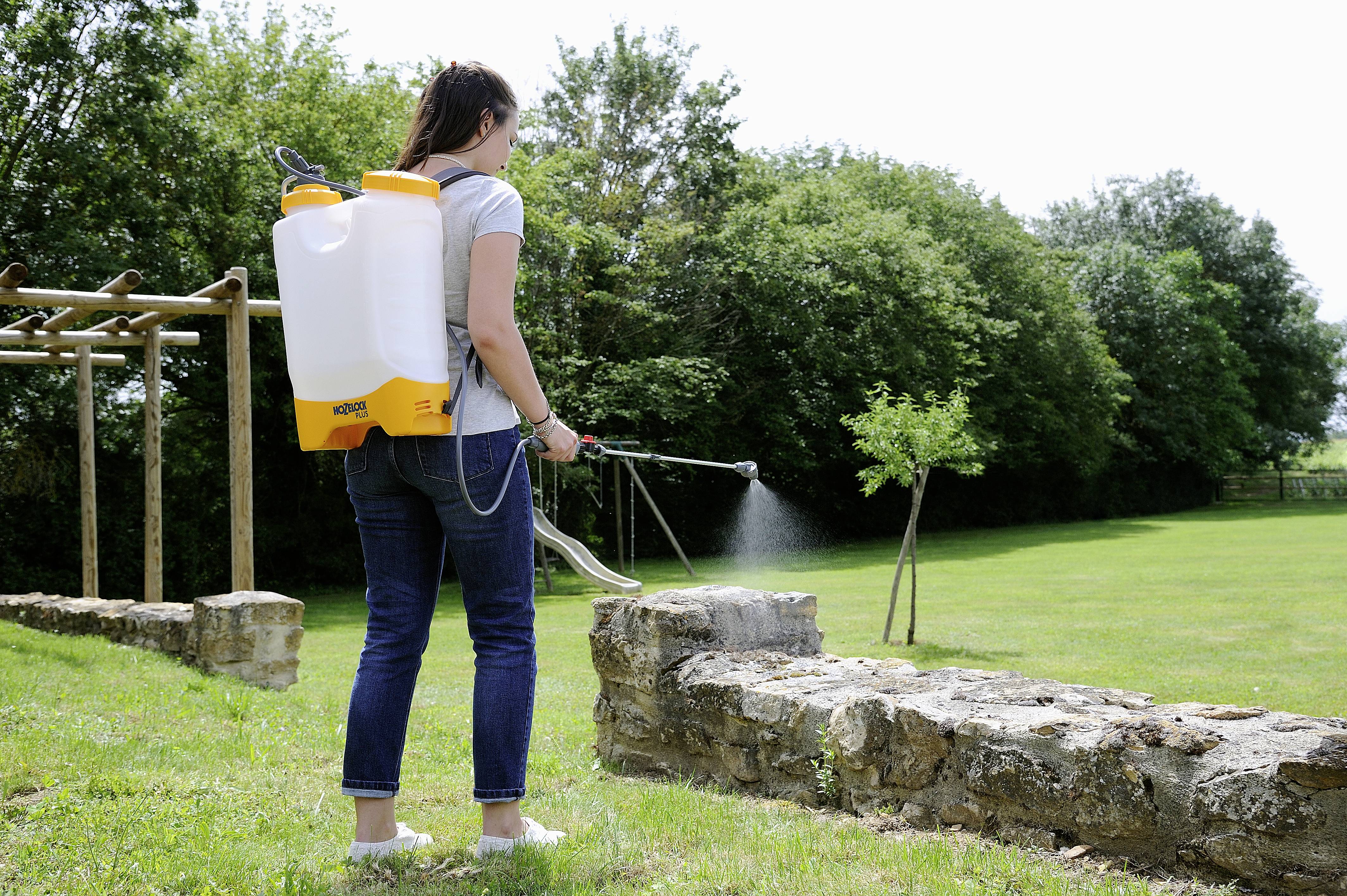 Eine Person sprüht Pflanzenschutzmittel aus einem Rucksack-Sprühgerät auf einen bepflanzten Bereich im Garten.