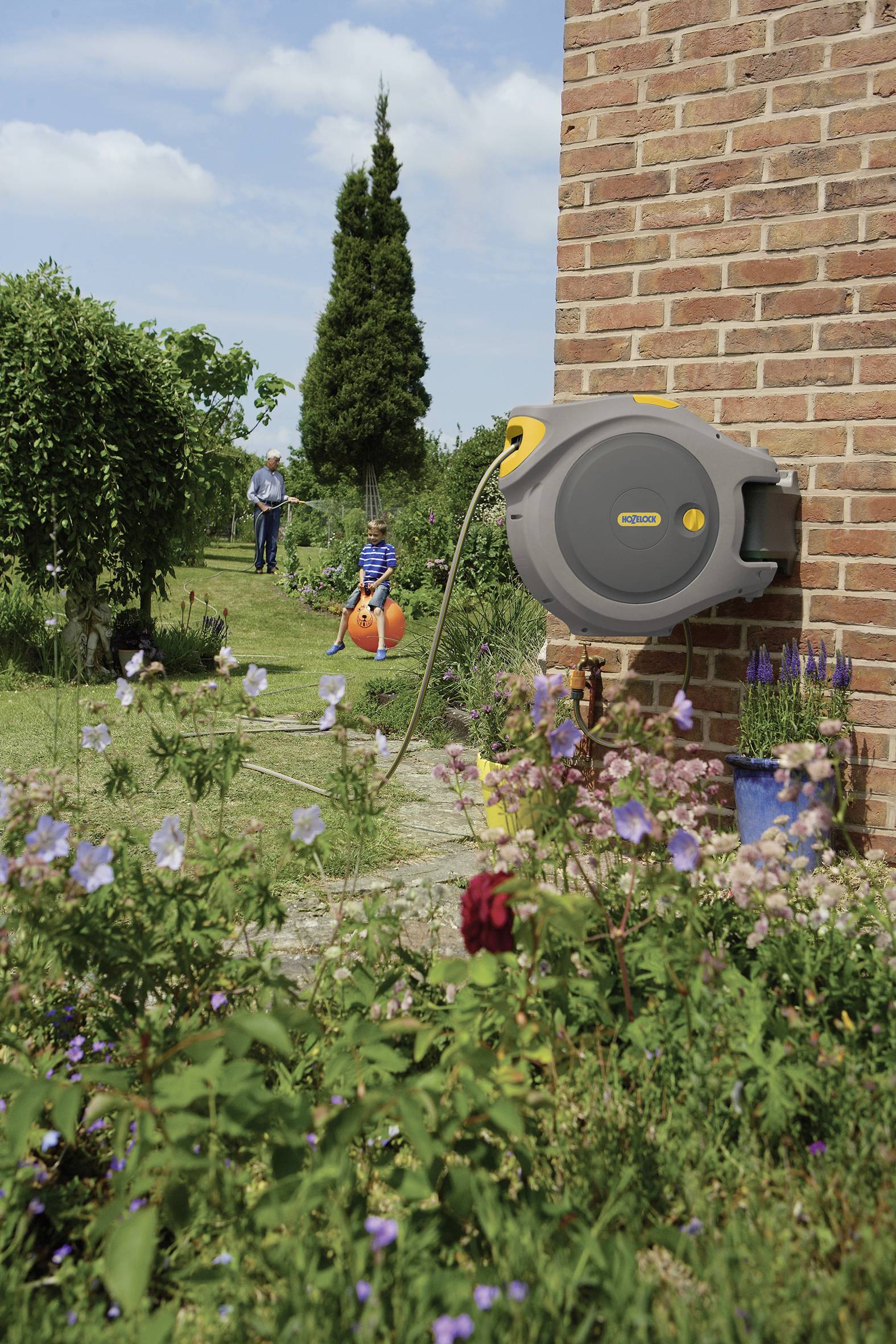 Eine Gartenszene mit einer aufgehängten Schlauchtrommel an einer Backsteinwand, im Hintergrund ein Kind auf einem Hüpfball und ein Erwachsener.