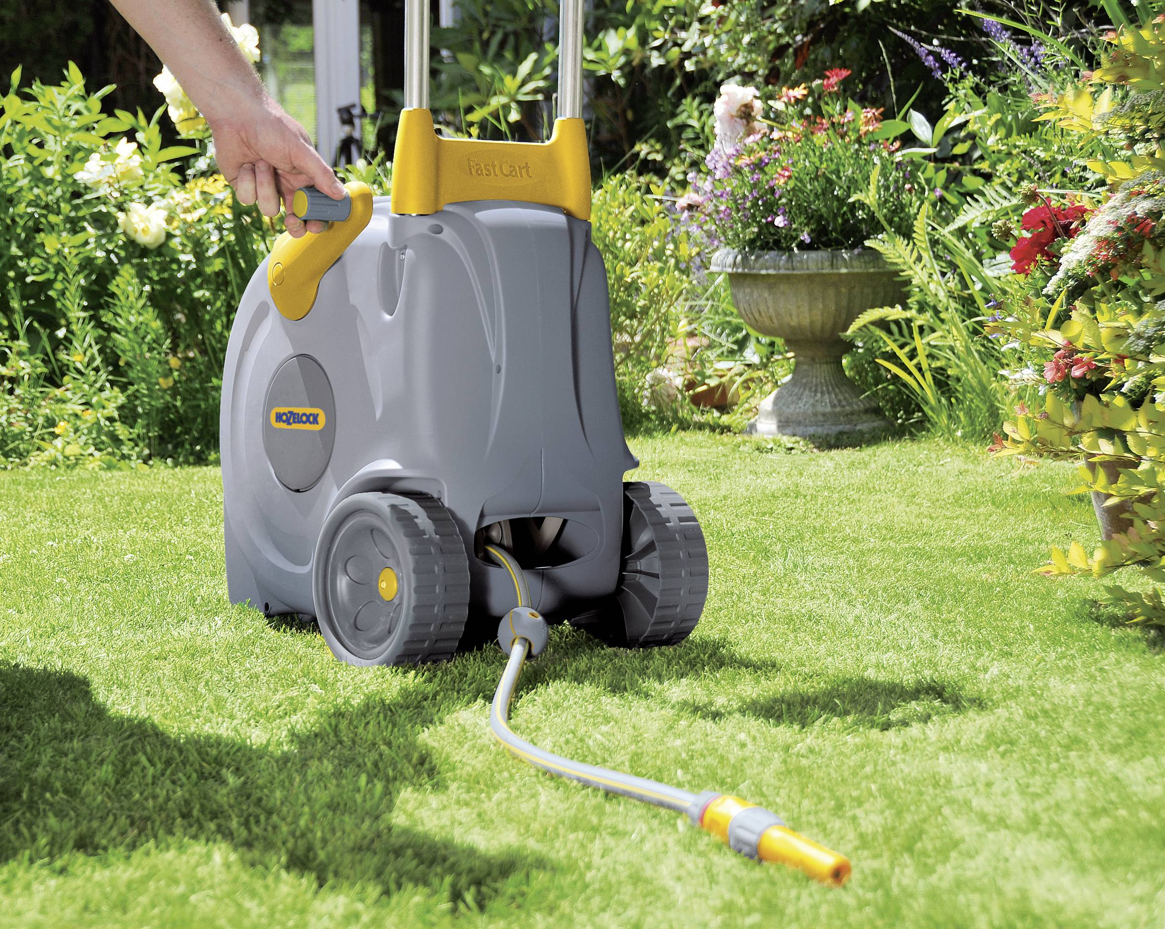 Eine Person zieht einen grauen Schlauchwagen mit gelben Details über eine grüne Rasenfläche in einem Garten mit Blumenbeeten.