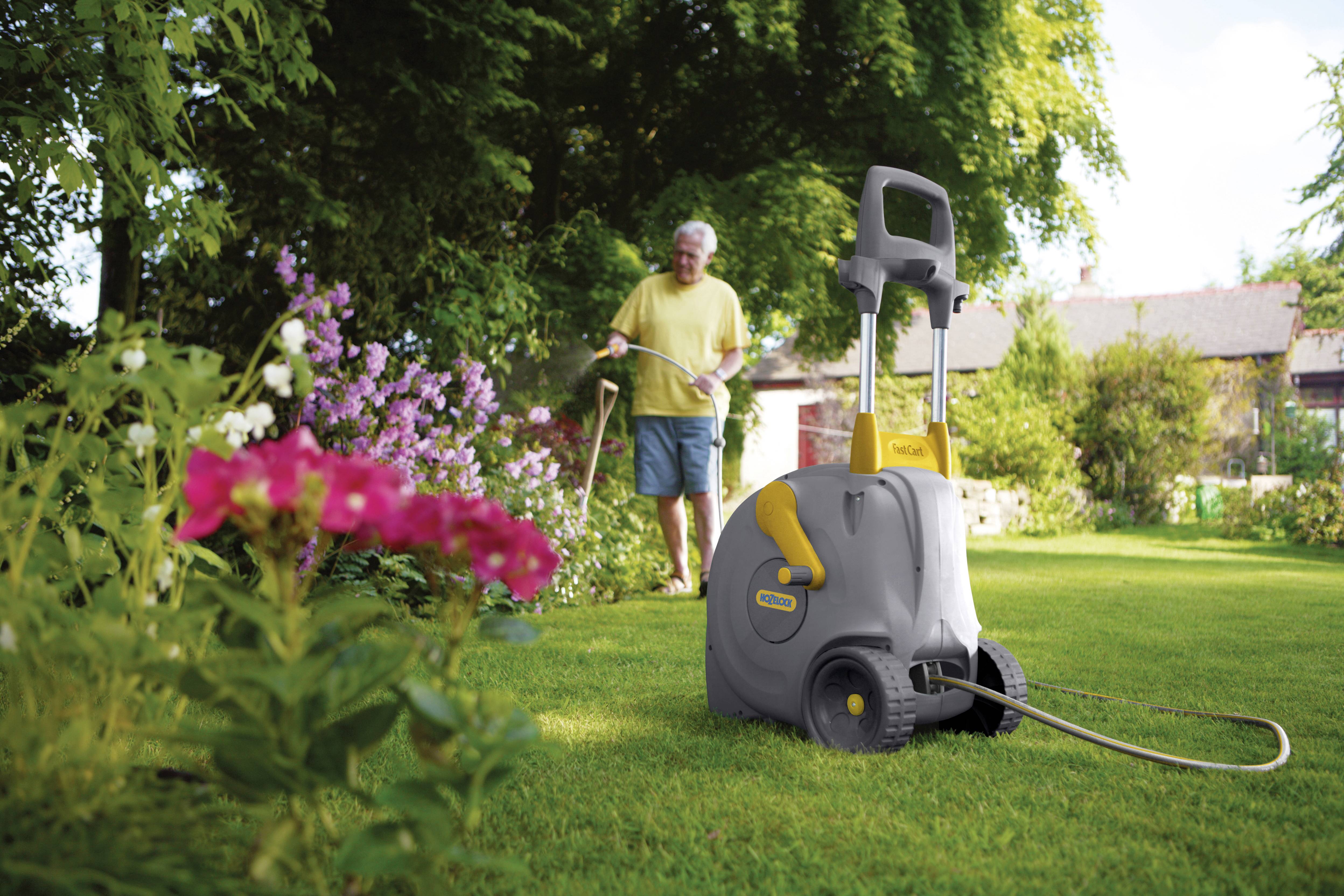 Ein Mann bewässert einen Garten mit einem Schlauchwagen auf einem sonnigen Rasen, umgeben von blühenden Blumen und Bäumen.