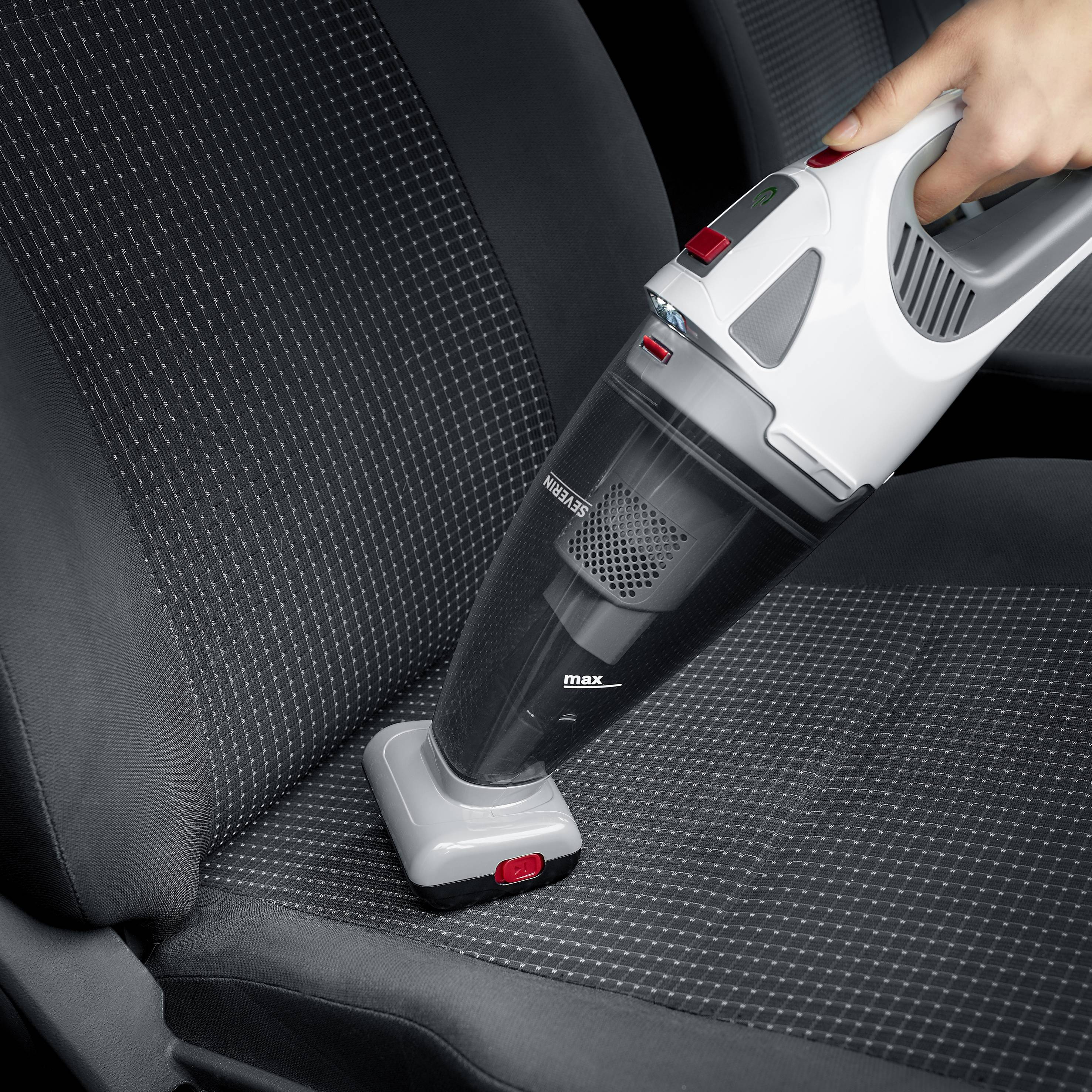 Eine Hand hält einen tragbaren Staubsauger, der den Sitz eines Autos reinigt. Der Staubsauger ist schwarz-grau mit einem roten Knopf.