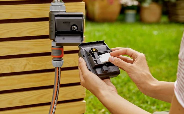 Person hält ein Gartengerät, während er eine Batterie einsetzt, verbunden mit einem Gartenschlauch vor einer Holzkulisse; Outdoor-Umgebung.