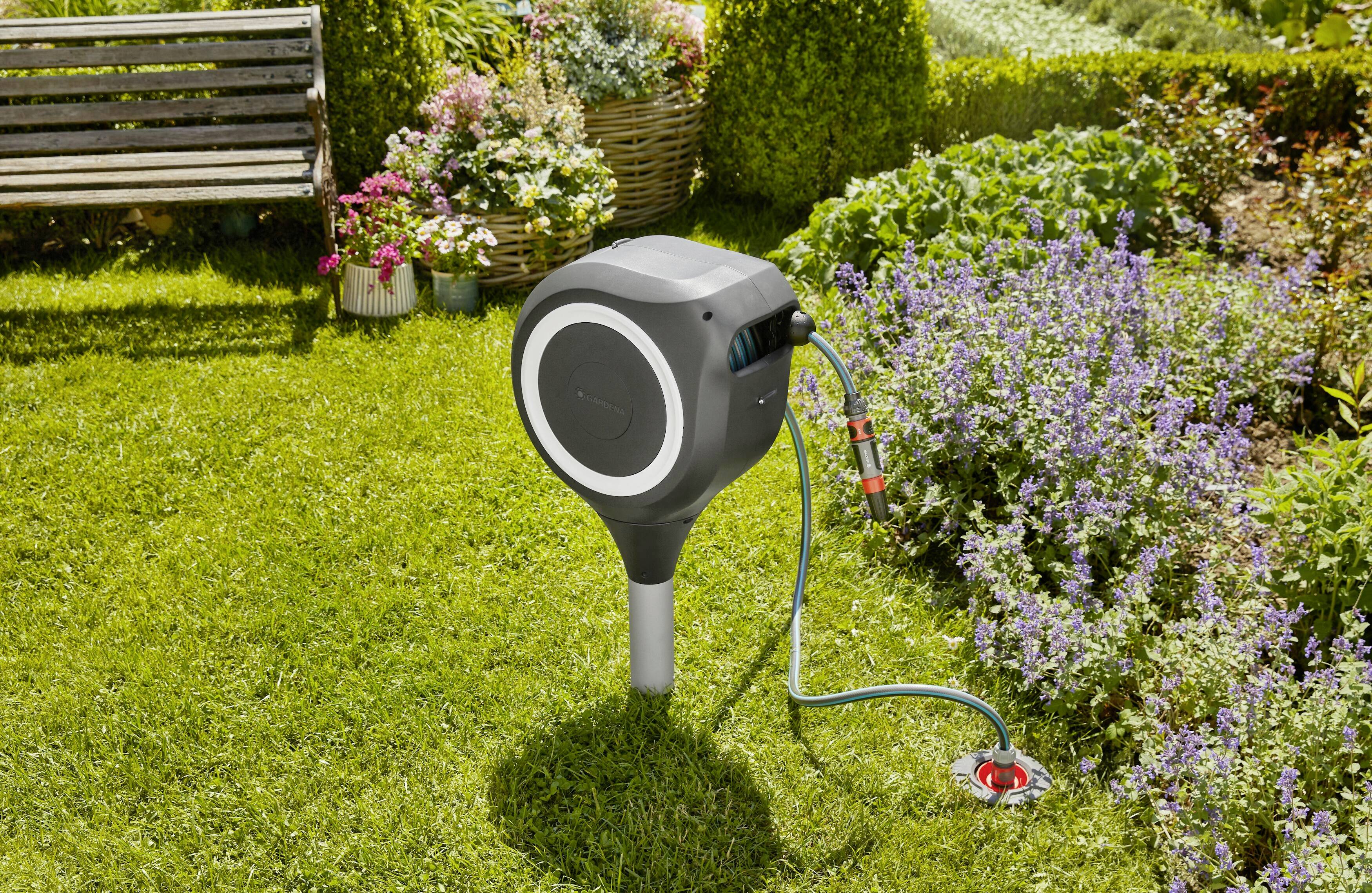 Eine aufgerollte Gartenschlauchbox steht im Grünen neben blühenden Pflanzen. Eine Bank und bunte Blumen sind im Hintergrund zu sehen.