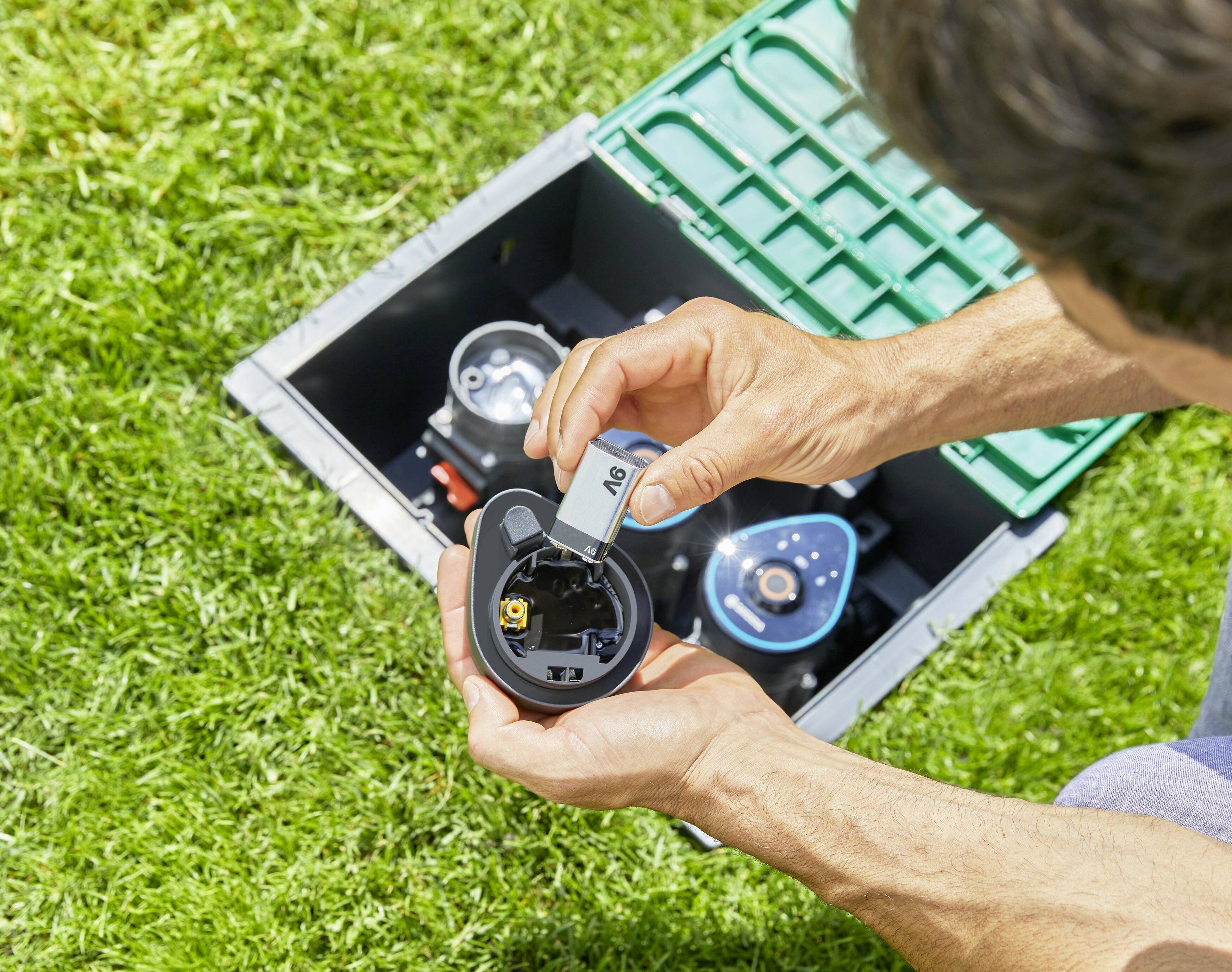 Eine Person installiert eine AA-Batterie in ein elektrisches Gerät im Freien, auf grünem Gras, neben einer schwarzen Box mit grünen Details.