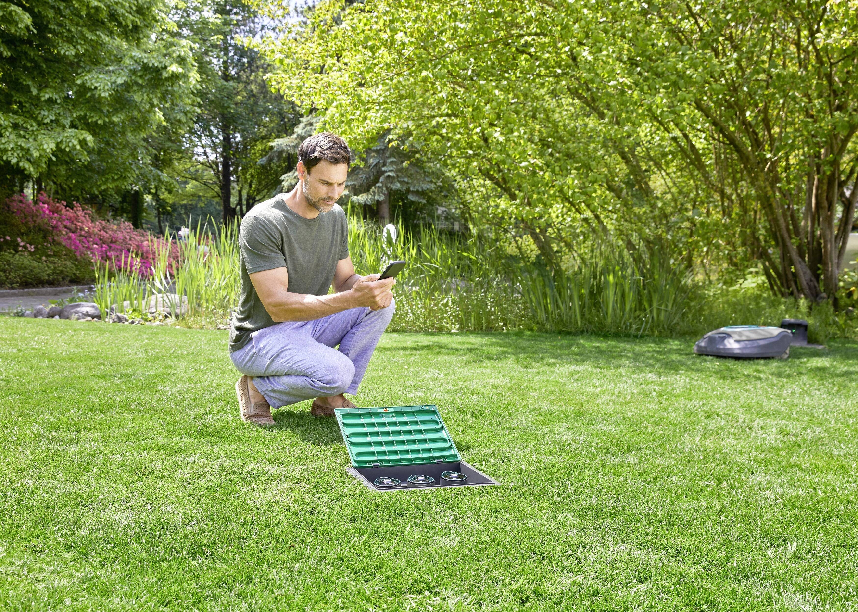 Ein Mann kniet auf einer Rasenfläche und bedient ein Bewässerungssystem über sein Smartphone. Im Hintergrund sind Bäume und Büsche.