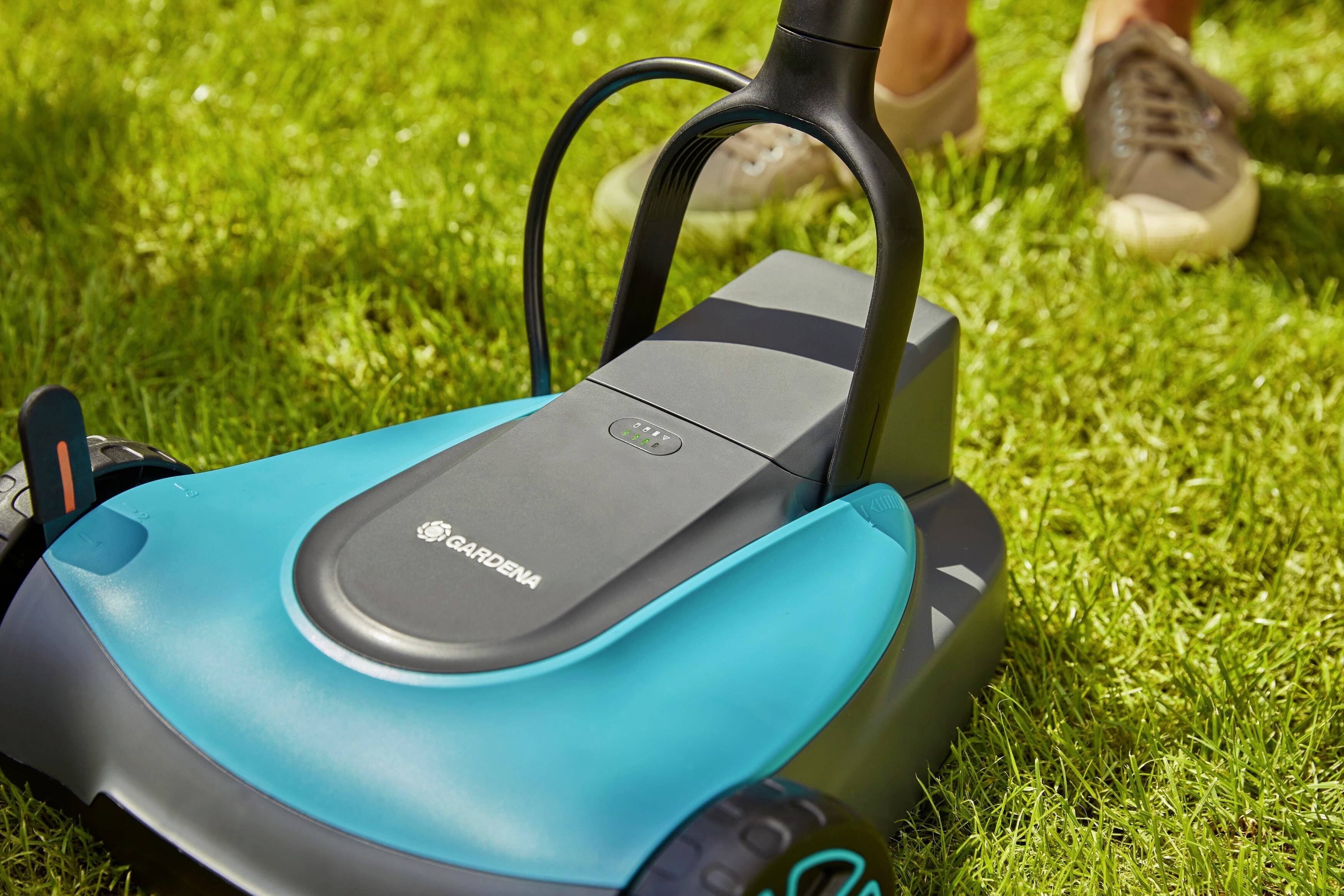 Ein blauer Rasenmäher von Gardena auf grünem Gras. Im Hintergrund sind unscharfe Beine und Schuhe einer Person zu sehen.