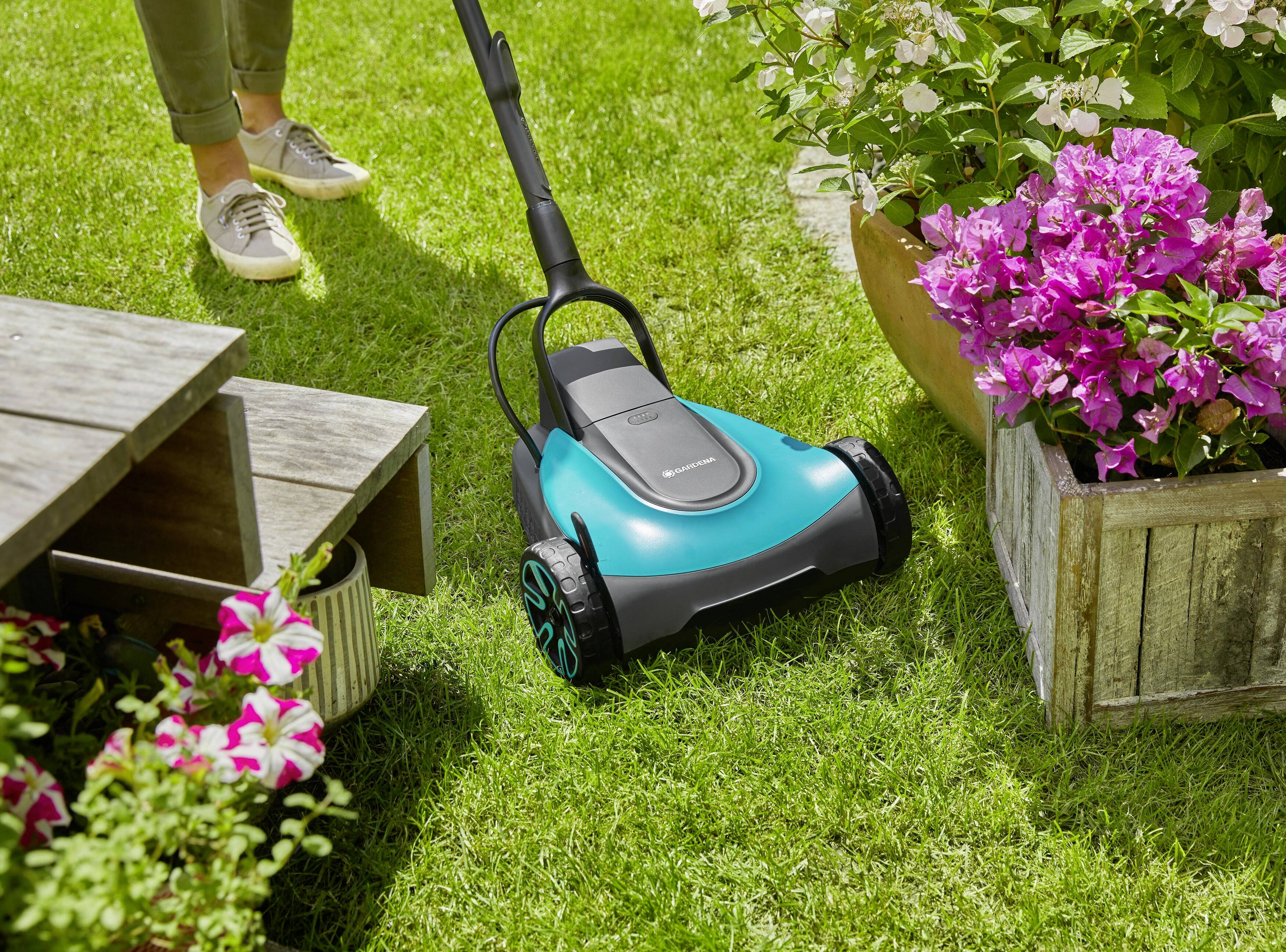 Eine Person mäht den Rasen mit einem blauen Rasenmäher neben blühenden Blumen und einer Holzterrasse.