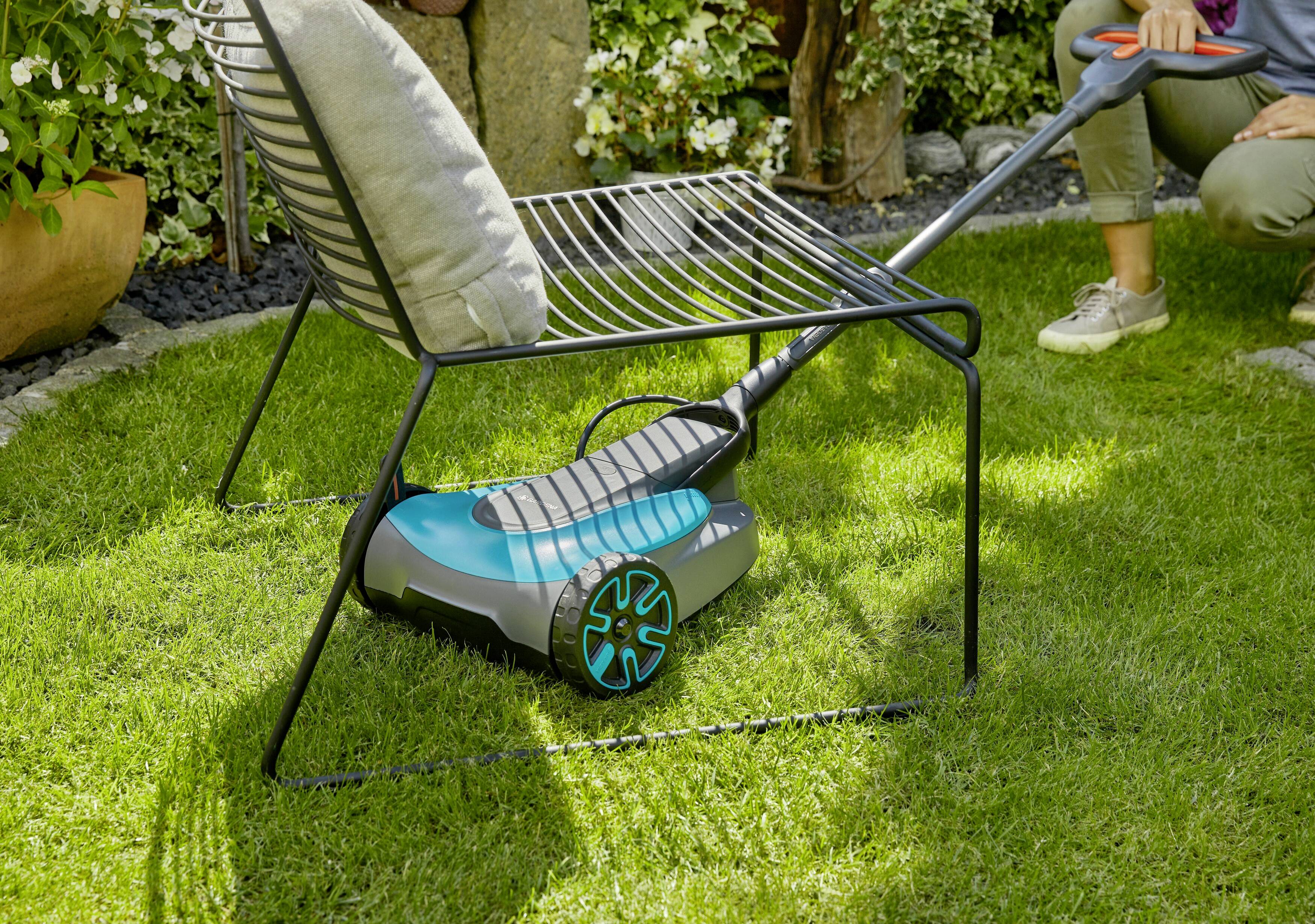 Person mäht Rasen in einem Garten mit einem kleinen, handgeführten Rasenmäher. Pflanzen und Gartenmöbel sind im Hintergrund zu sehen.