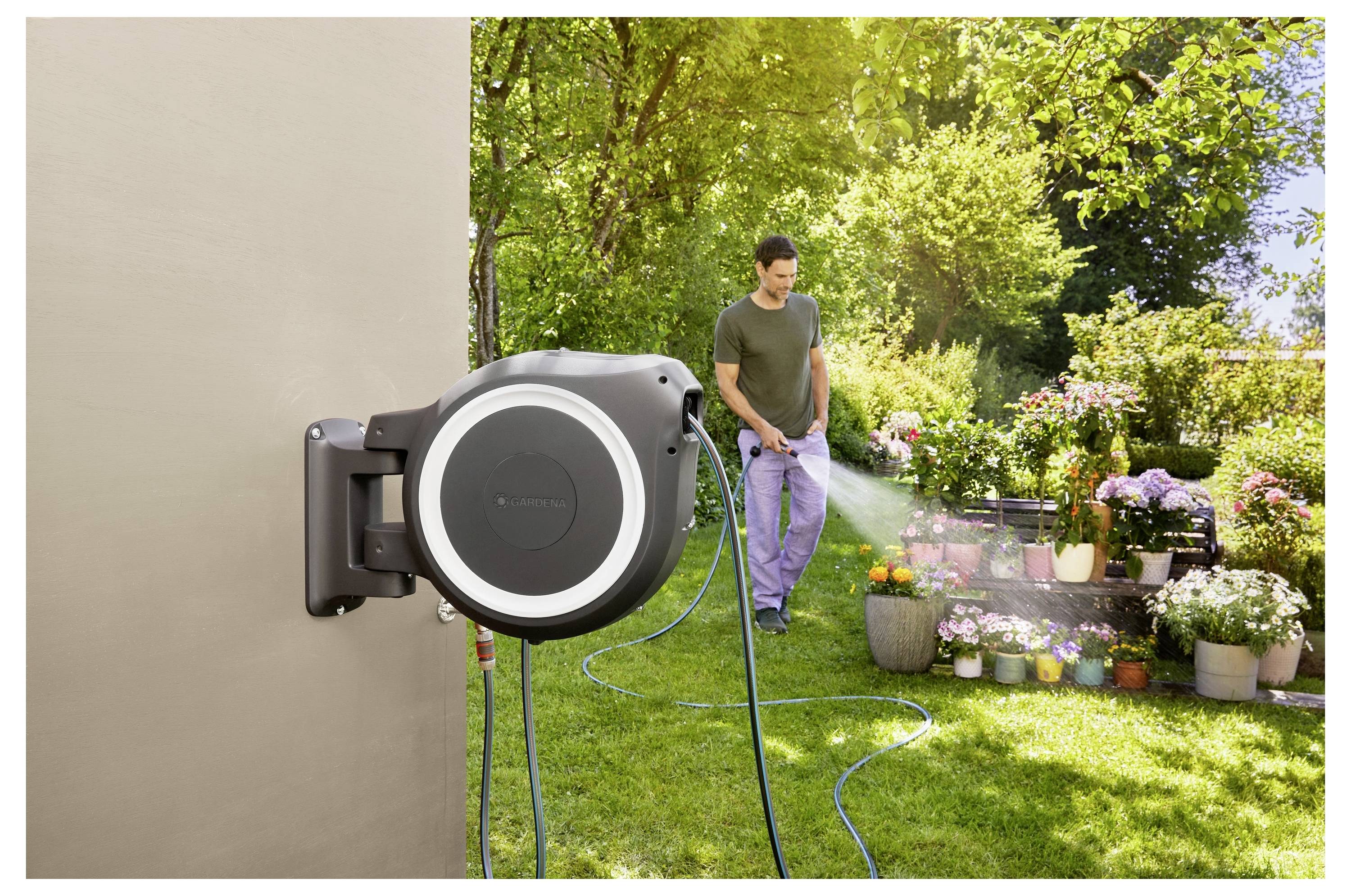 Eine Person bewässert einen Garten mit einem Schlauch, der an einer wandmontierten Schlauchtrommel angeschlossen ist. Sie sind von Pflanzen und Blumen in Töpfen umgeben.