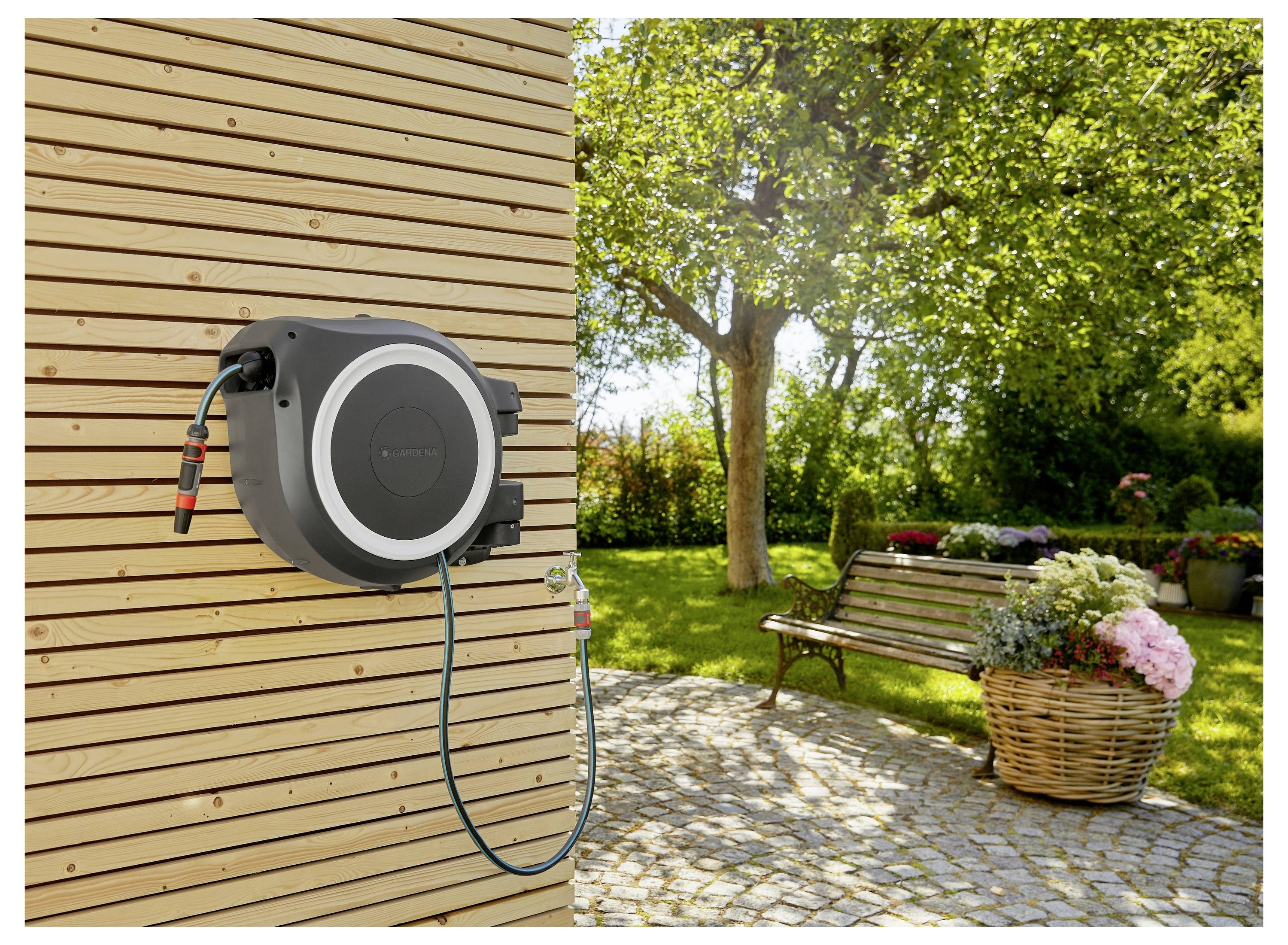Eine ausziehbare Gartensch­lauchrolle, die an einer Holzwand in der Nähe einer Terrasse montiert ist, mit einem Blumenkorb und einer Bank. Ein sonniger Garten mit Bäumen.