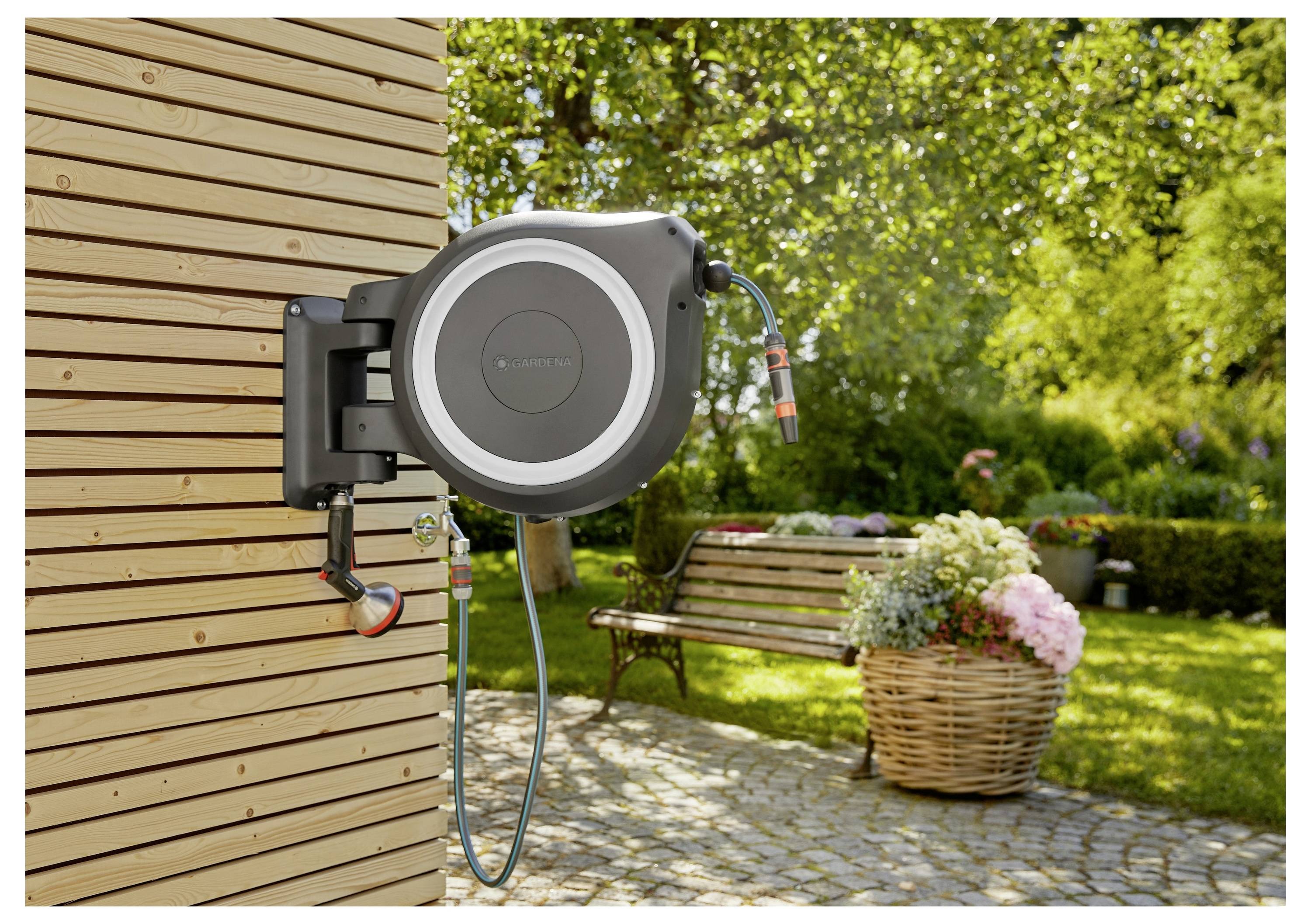 Eine an der Wand montierte Gartenschlauchrolle ist an einer Holzwand befestigt, mit einem Schlauch, der darunter verläuft. Ein Garten mit einer Bank und einem Blumenkorb erscheint im Hintergrund.