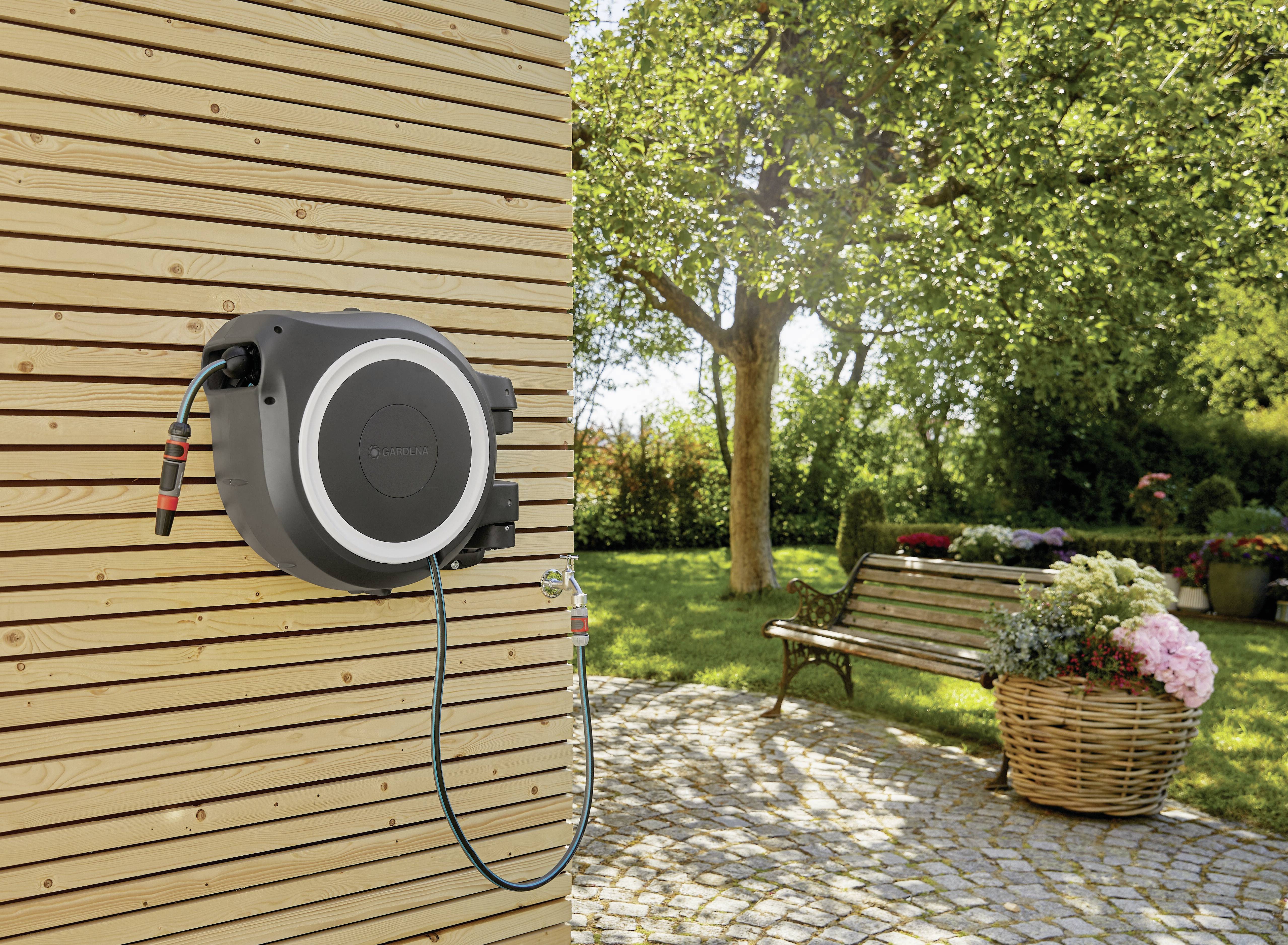 Ein automatischer Gartenschlauchaufroller ist an einer Holzwand montiert. Im Hintergrund ist ein grüner Garten mit Bäumen und Blumen zu sehen.