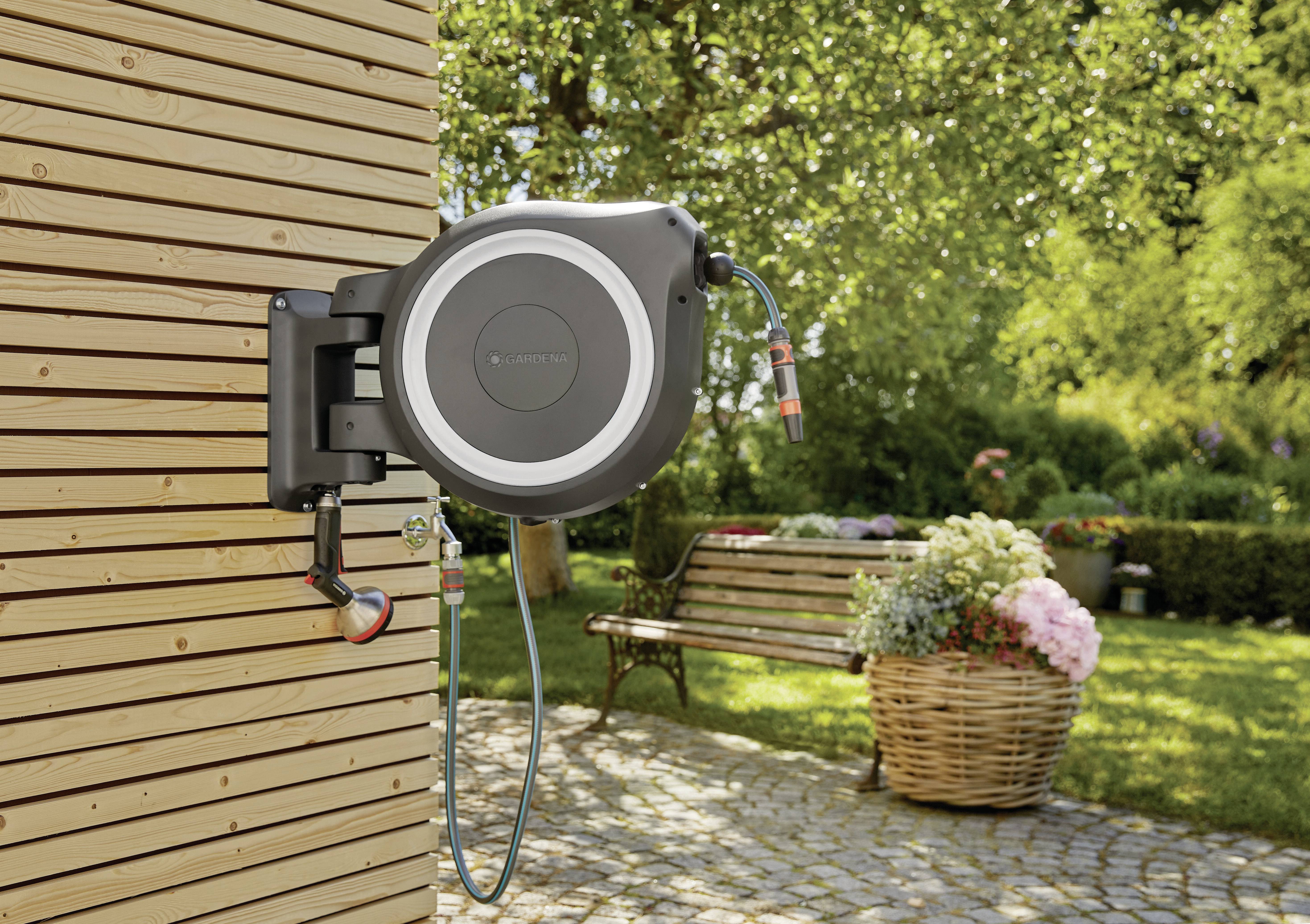 Eine automatische Gartenschlauchtrommel ist an einer Holzwand montiert. Im Hintergrund befindet sich ein Garten mit einer Bank und Blumen.
