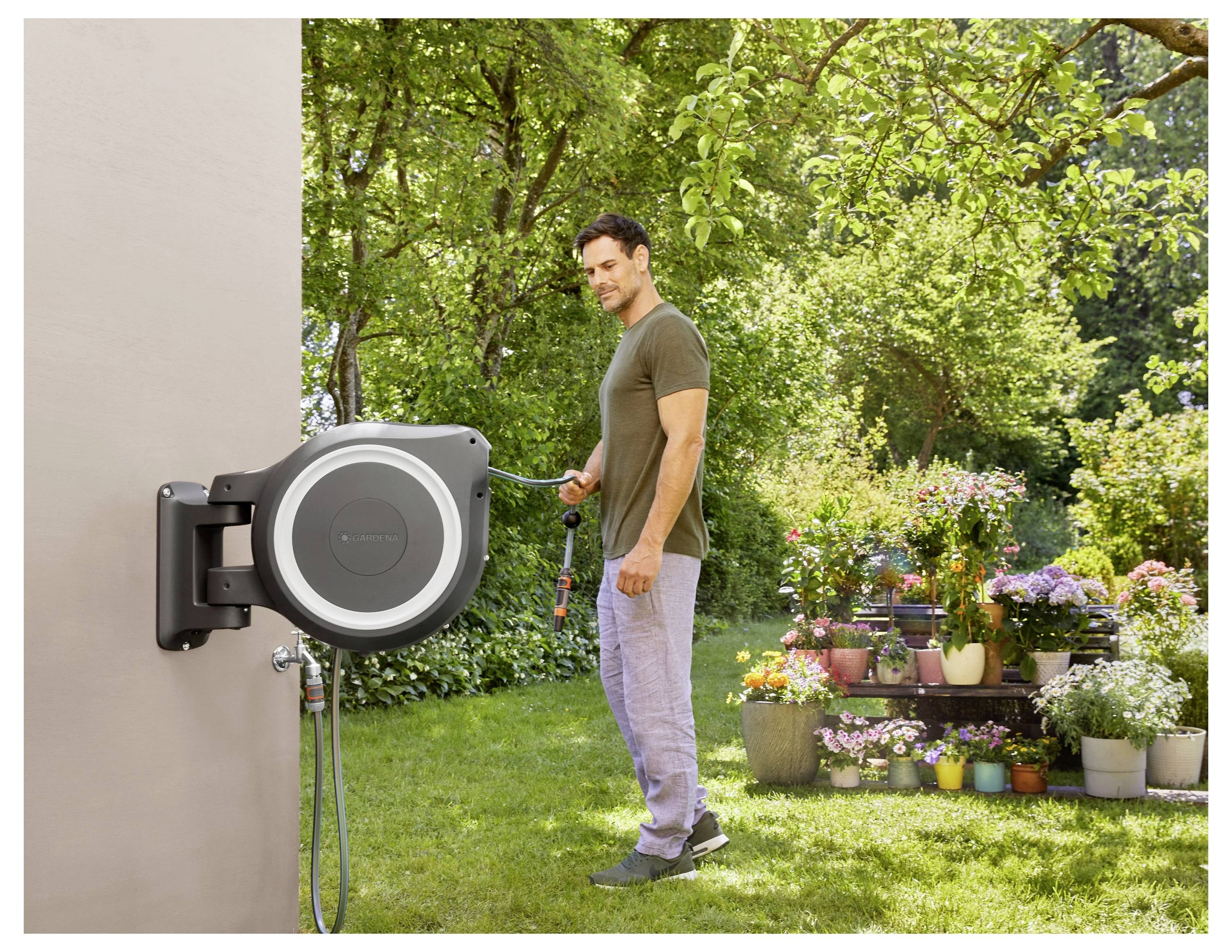 Eine Person wickelt eine an der Wand befestigte Gartenschlauchrolle ab, umgeben von verschiedenen Blütenpflanzen und Bäumen, an einem sonnigen Tag.