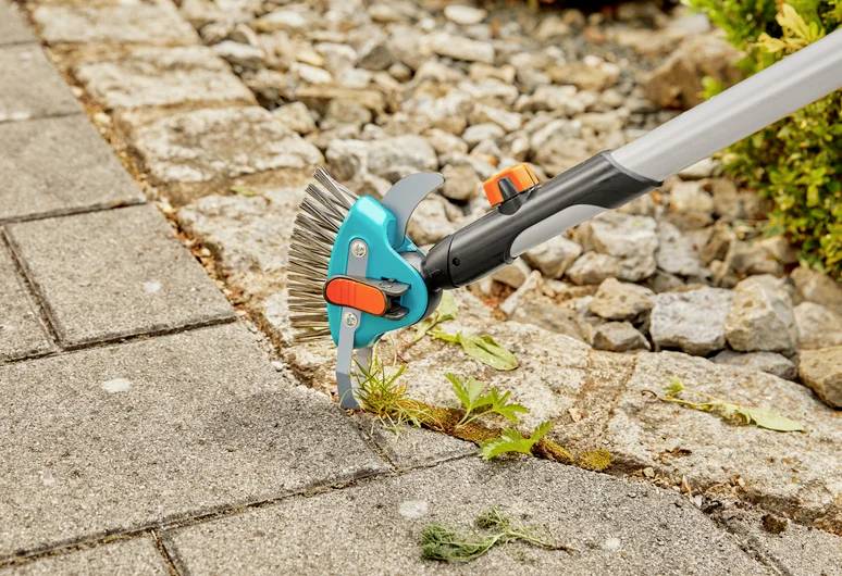 Eine Gartenhacke entfernt Unkraut zwischen Pflastersteinen und zeigt eine Nahaufnahme ihrer Anwendung auf einem Weg, der von Steinen und Grünpflanzen umgeben ist.